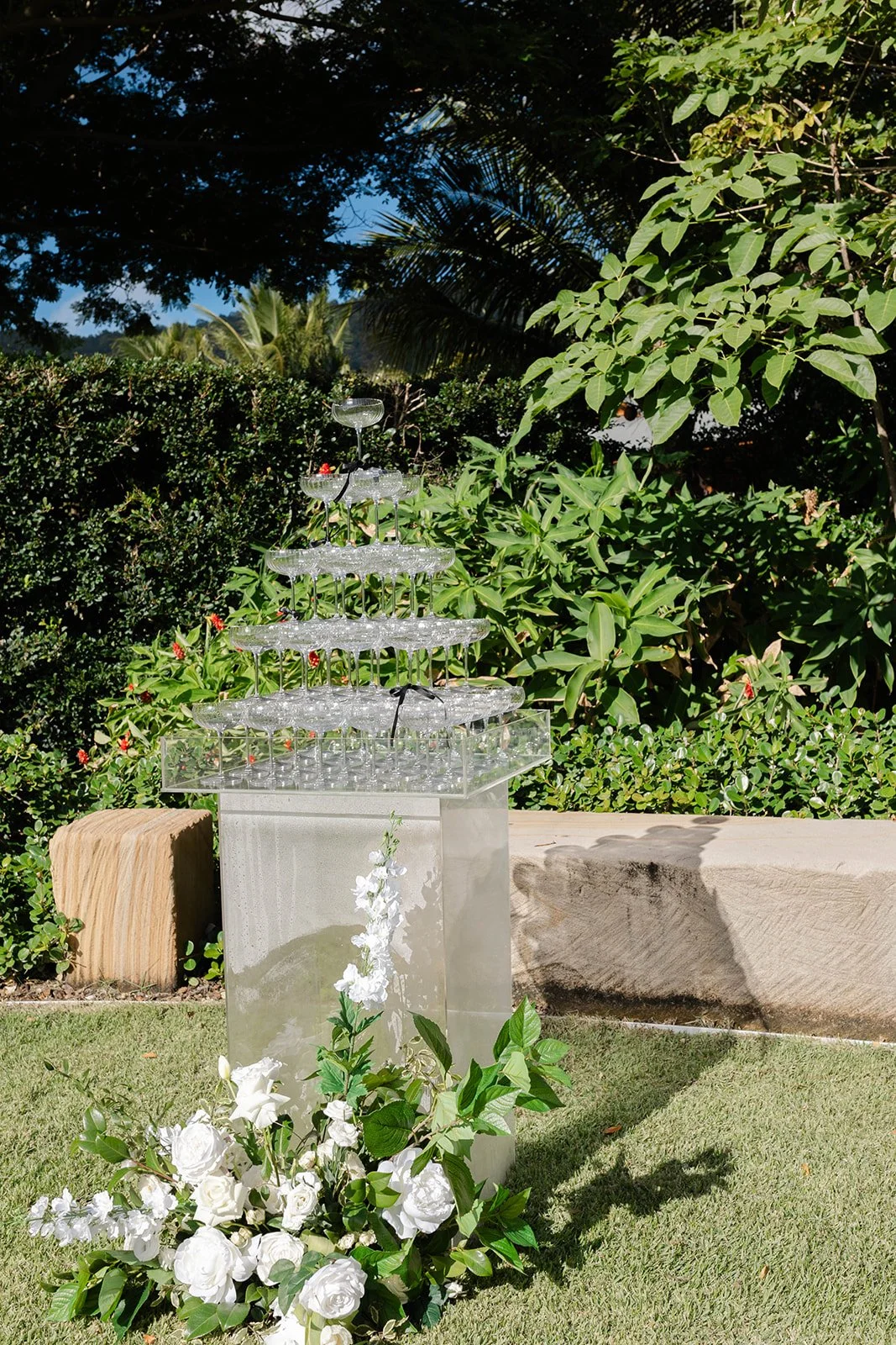 A champagne tower made of stacked glasses on a transparent stand, decorated with a white floral arrangement, set outdoors with green bushes and trees in the background.