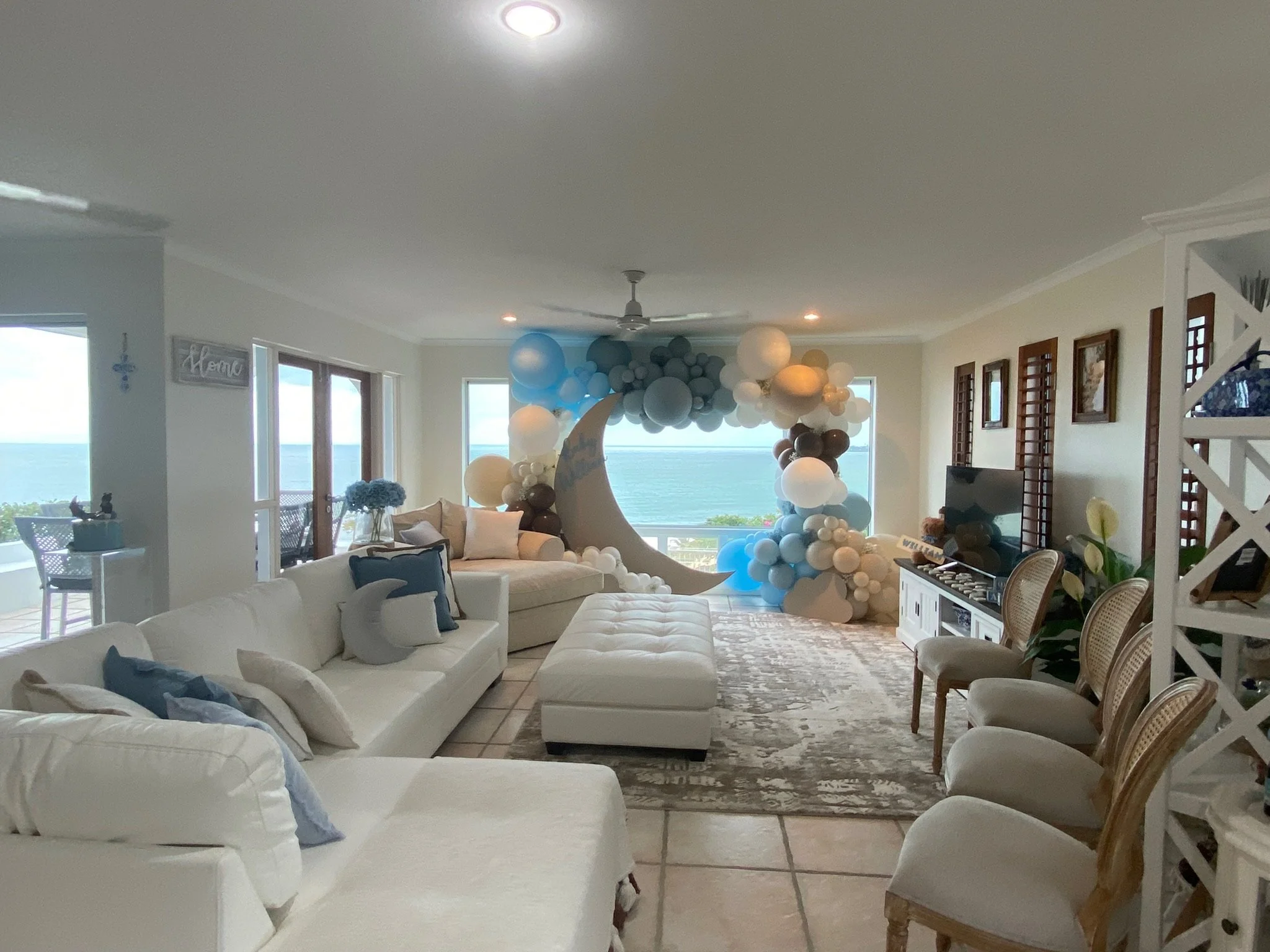 Living room decorated with balloons in shades of blue, white, and brown, featuring a large crescent moon and cloud cutouts, with ocean view through windows, white sofas, an ottoman, a flat-screen TV, and chairs.