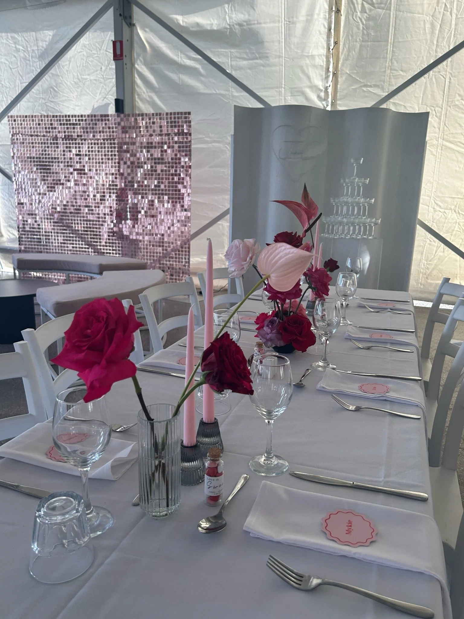 Elegant table setup with white tablecloth, pink and red floral centerpieces, pink candles, and place cards in a bright tent.