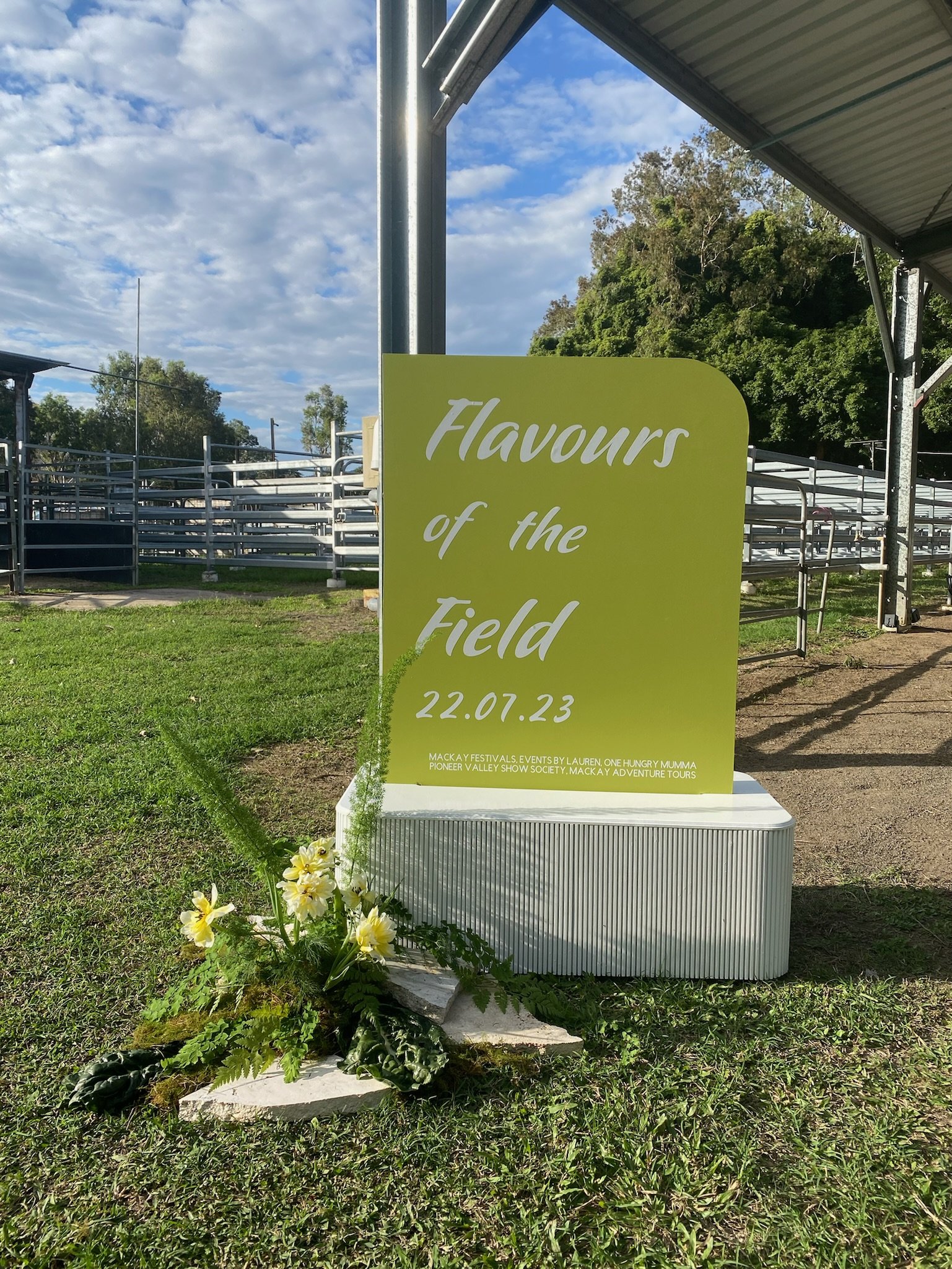 A sign that reads 'Flavours of the Field 22.07.23' on a grassy area, with a small flower arrangement at the base. The background includes a goal post, a covered structure, and trees under a partly cloudy sky.