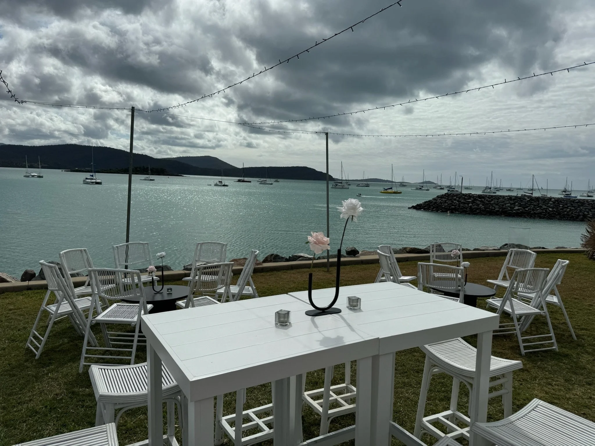 Outdoor patio setup with white tables and chairs overlooking a body of water with sailboats, cloudy sky, and distant hills.