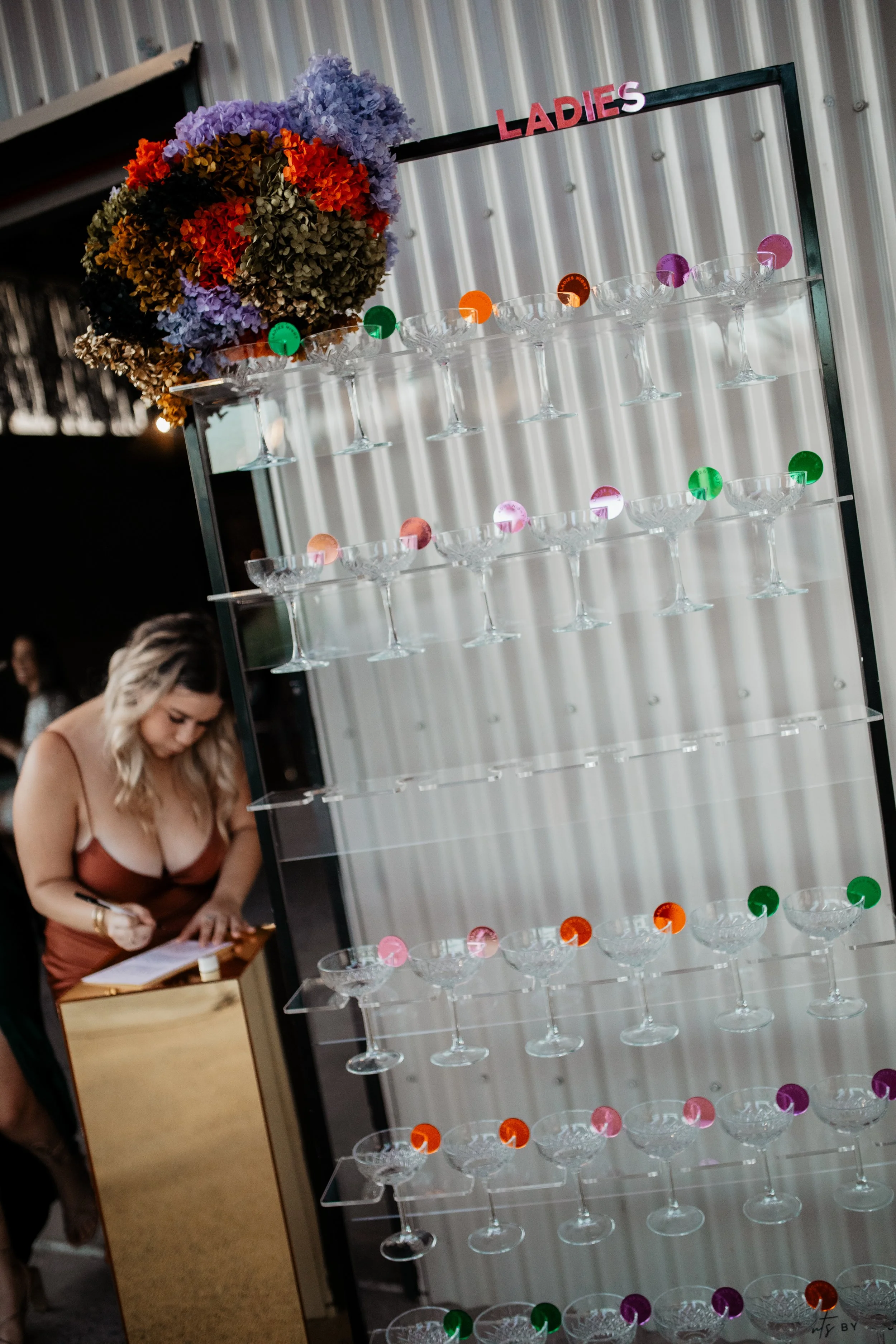 A display of empty cocktail glasses with colored tabs on top, labeled 'Ladies', with a person taking notes in the background.