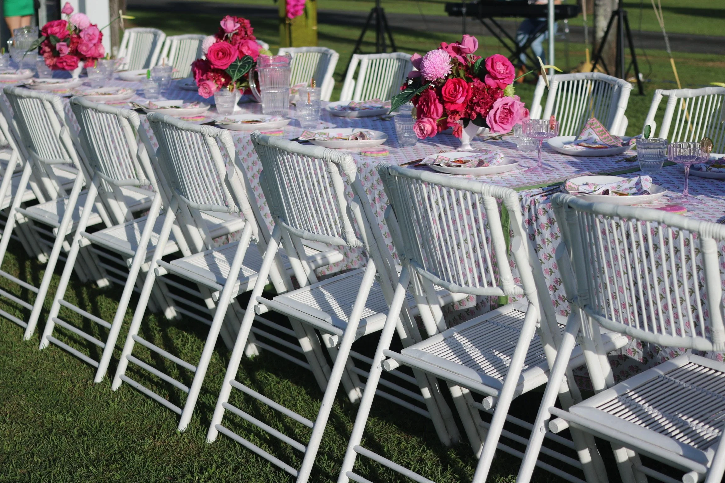 Outdoor event table decorated with pink and purple floral centerpieces, set with plates, glassware, and napkins on a floral tablecloth, surrounded by white chairs on grass.