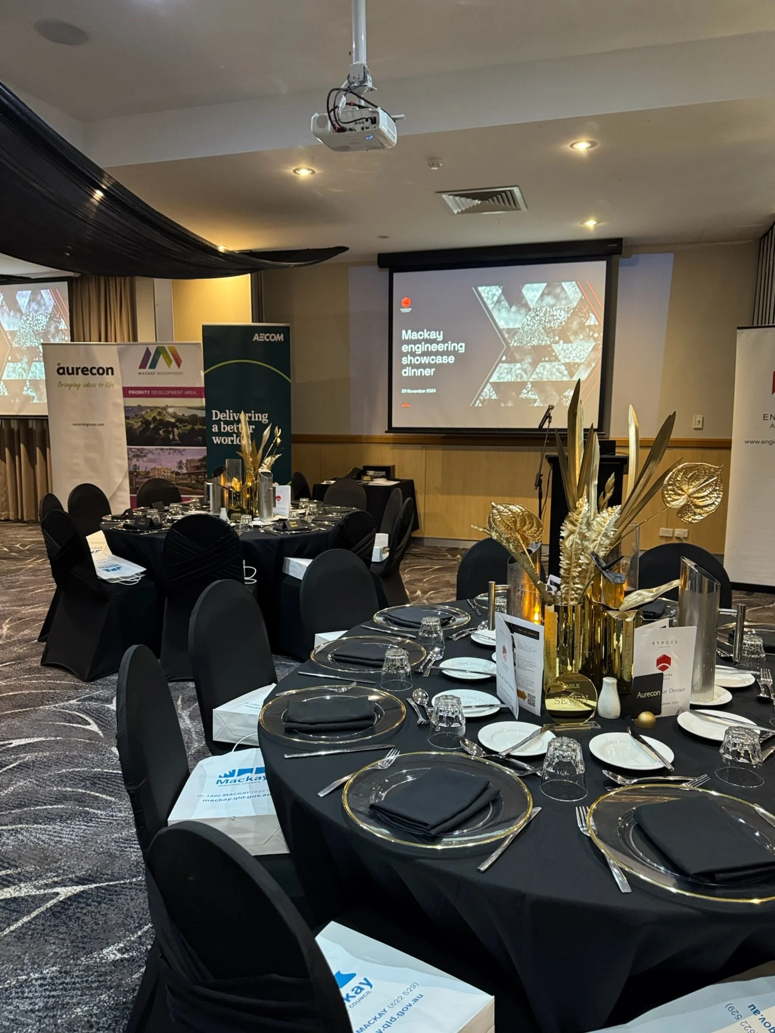 A banquet hall set for an event with round tables covered in black tablecloths, decorated with gold and white centerpieces, black napkins, and glassware. A large screen displays a presentation titled 'Mackay engineering showcase dinner.' There are ba