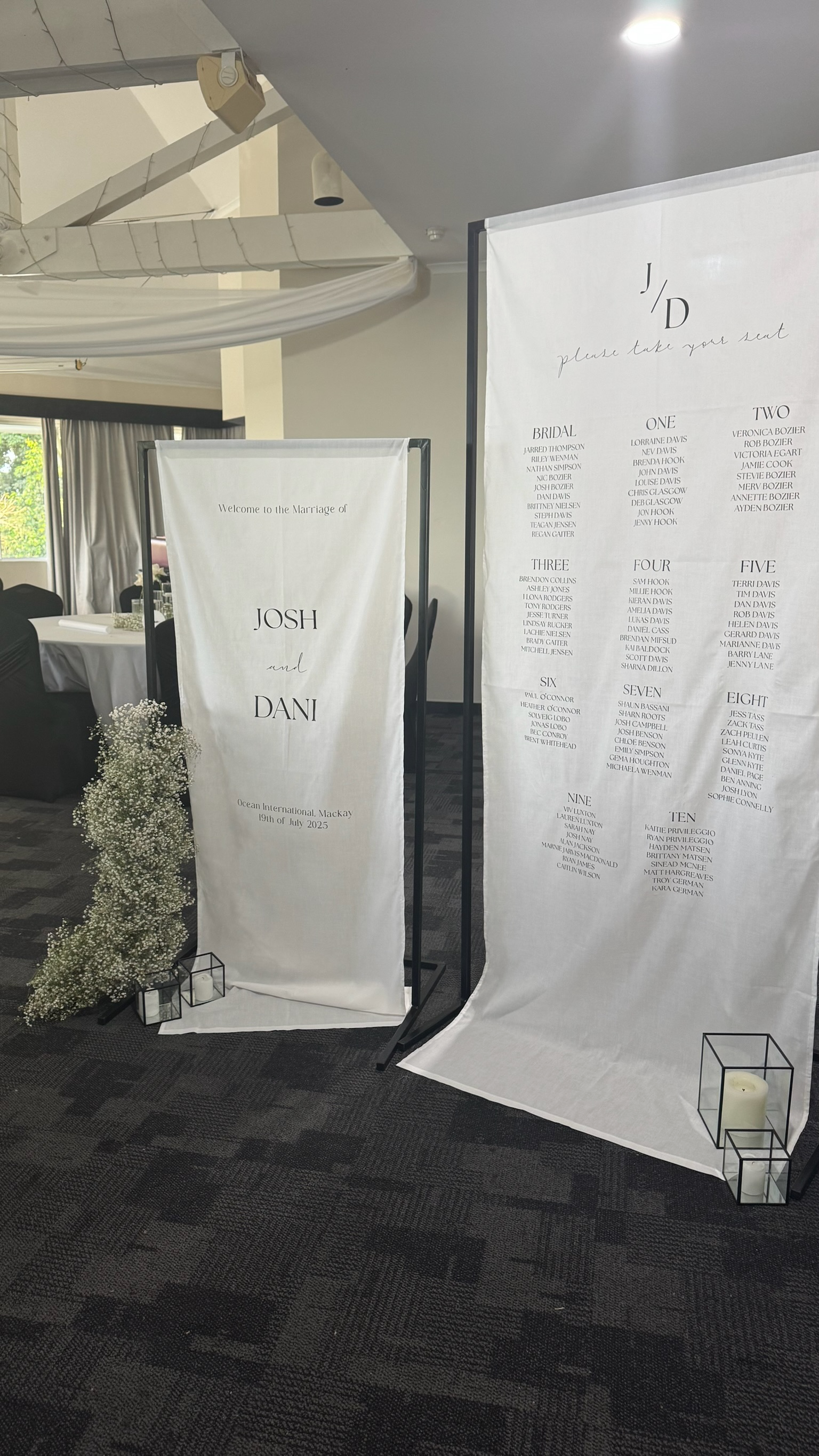 Wedding seating plan and welcome sign at a wedding reception with white fabric banners and black metal frames, flowers, and candles.