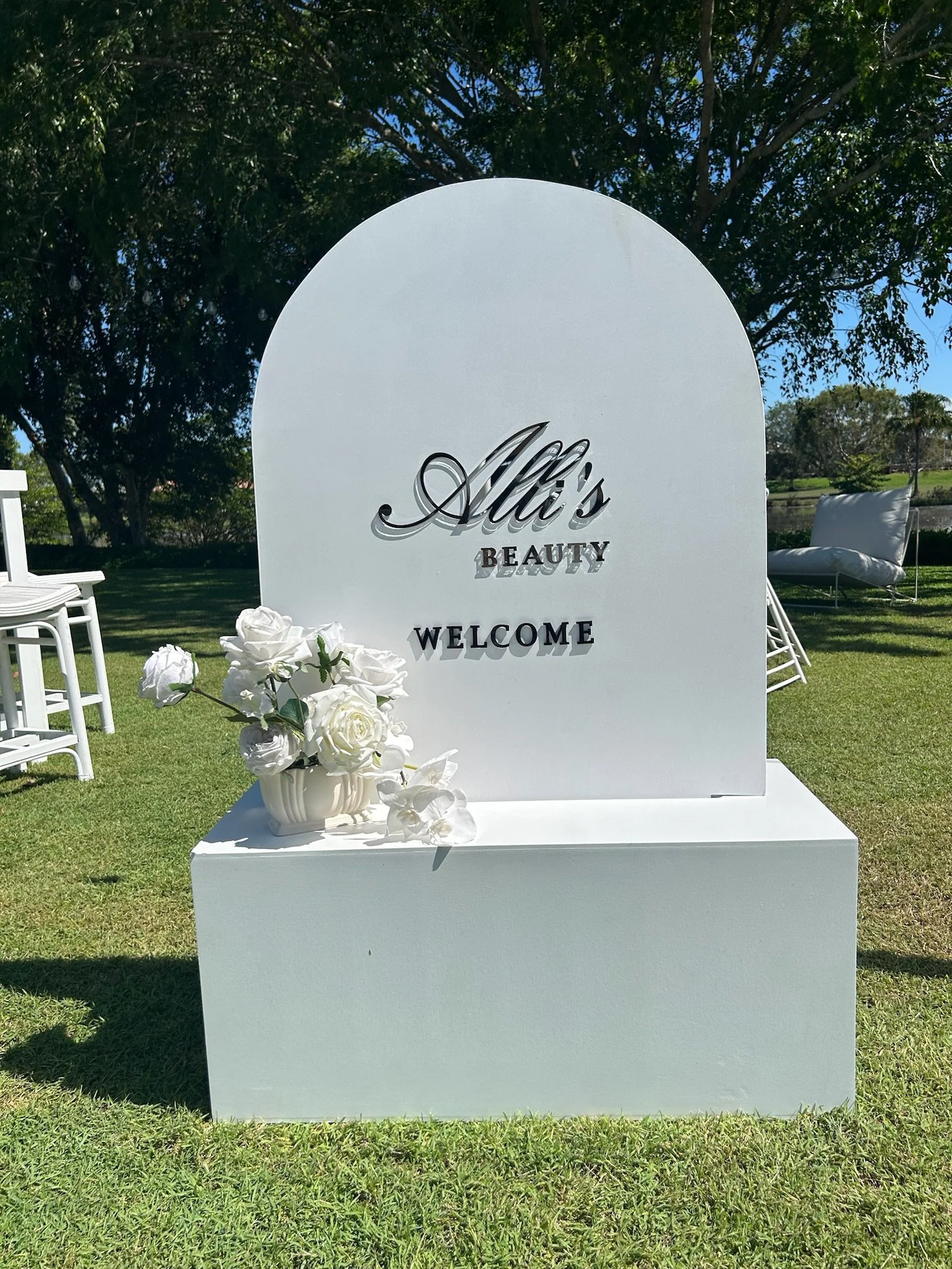 White outdoor sign with black lettering reading 'Ali's Beauty Welcome,' decorated with a white vase of white flowers, set on grass with trees and outdoor furniture in the background.