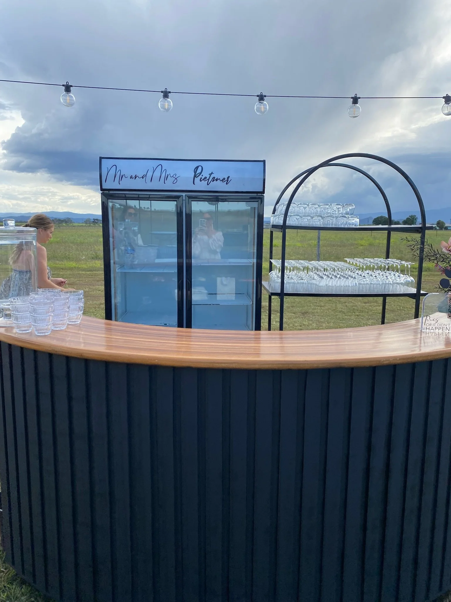 Outdoor bar setup with a black curved bar counter, glassware on a metal rack, a blue refrigerator with a sign reading "Mr and Mrs Pietner," and a woman in a floral dress, with string lights and an open field under a cloudy sky in the background.