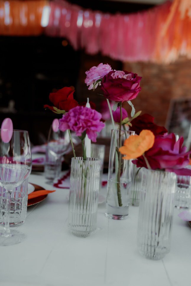 Table with pink, red, and orange flowers in clear glass vases, wine glasses, and place settings at a decorated event.