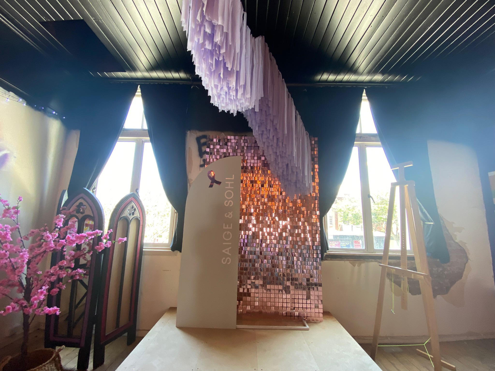 Indoor room with black drapes, pink decorative hanging installation, and a display with the logo and name 'Saige & SOHL.' There is a pink cherry blossom tree on the left, an easel on the right, and windows with daylight. There are some gold and pink 