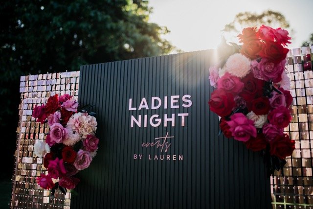 A sign reading 'Ladies Night' with floral decorations on each side and a sparkly backdrop, set outdoors during sunset.