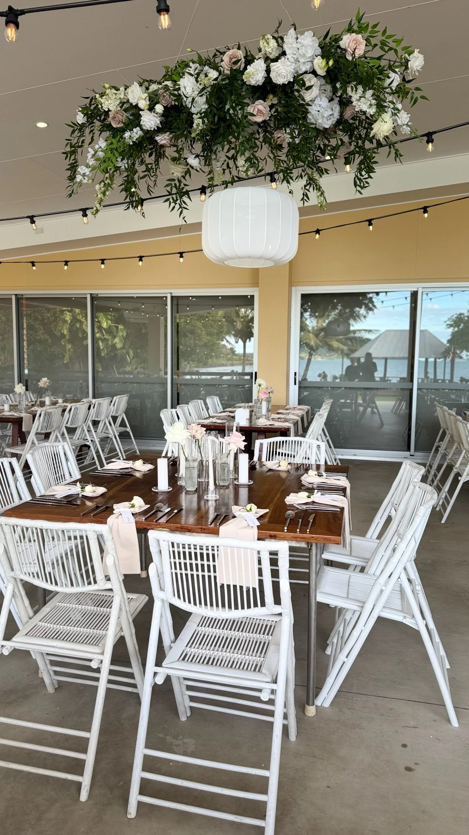 A decorated outdoor or semi-outdoor event space with white chairs, a large wooden table set for a meal, floral centerpieces, and a floral arrangement hanging from the ceiling. The background shows a view of trees and water through glass doors.