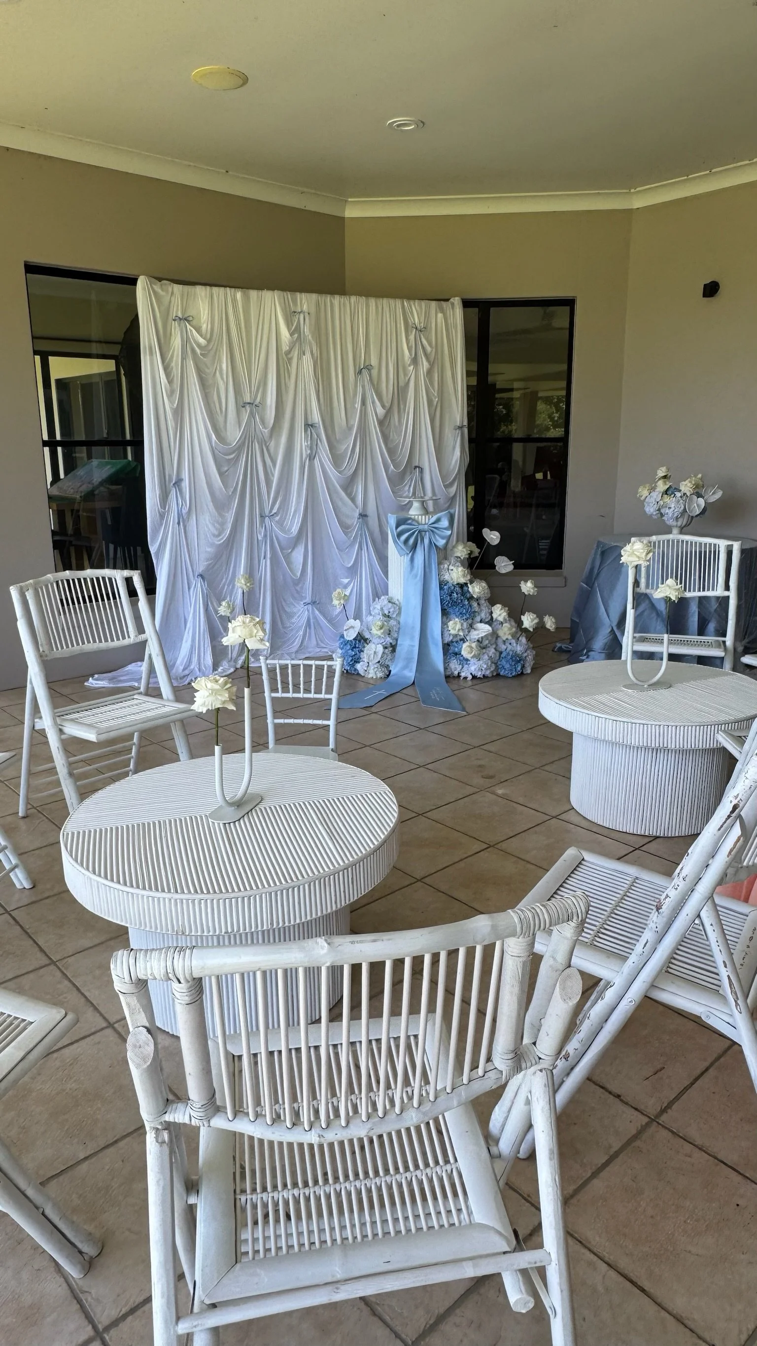 Wedding ceremony setup with white chairs, round tables with floral centerpieces, and a white curtain backdrop with blue accents and floral arrangements.