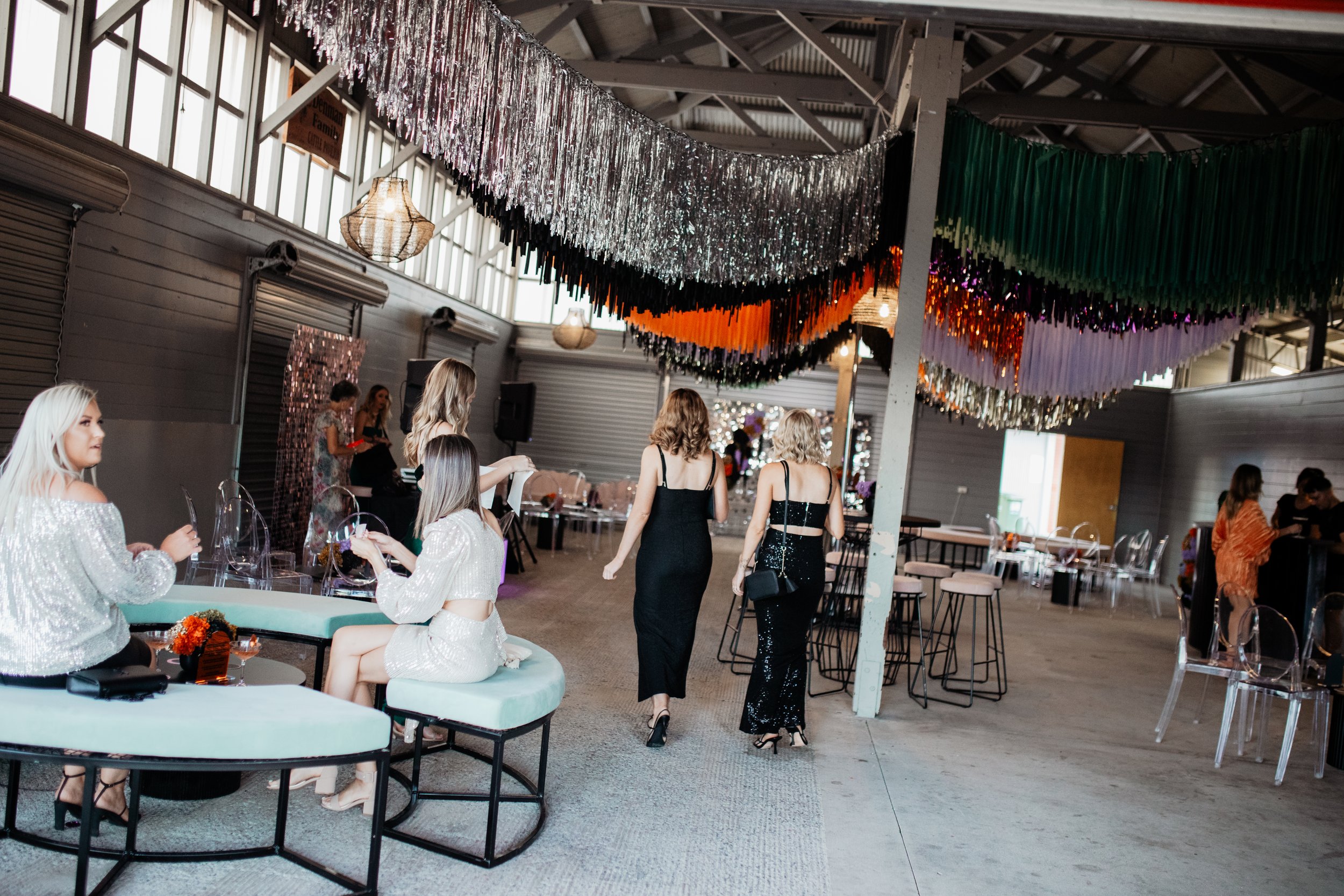 Indoor party venue decorated with colorful paper streamers and tinsel, with women in elegant dresses mingling and sitting at tables.