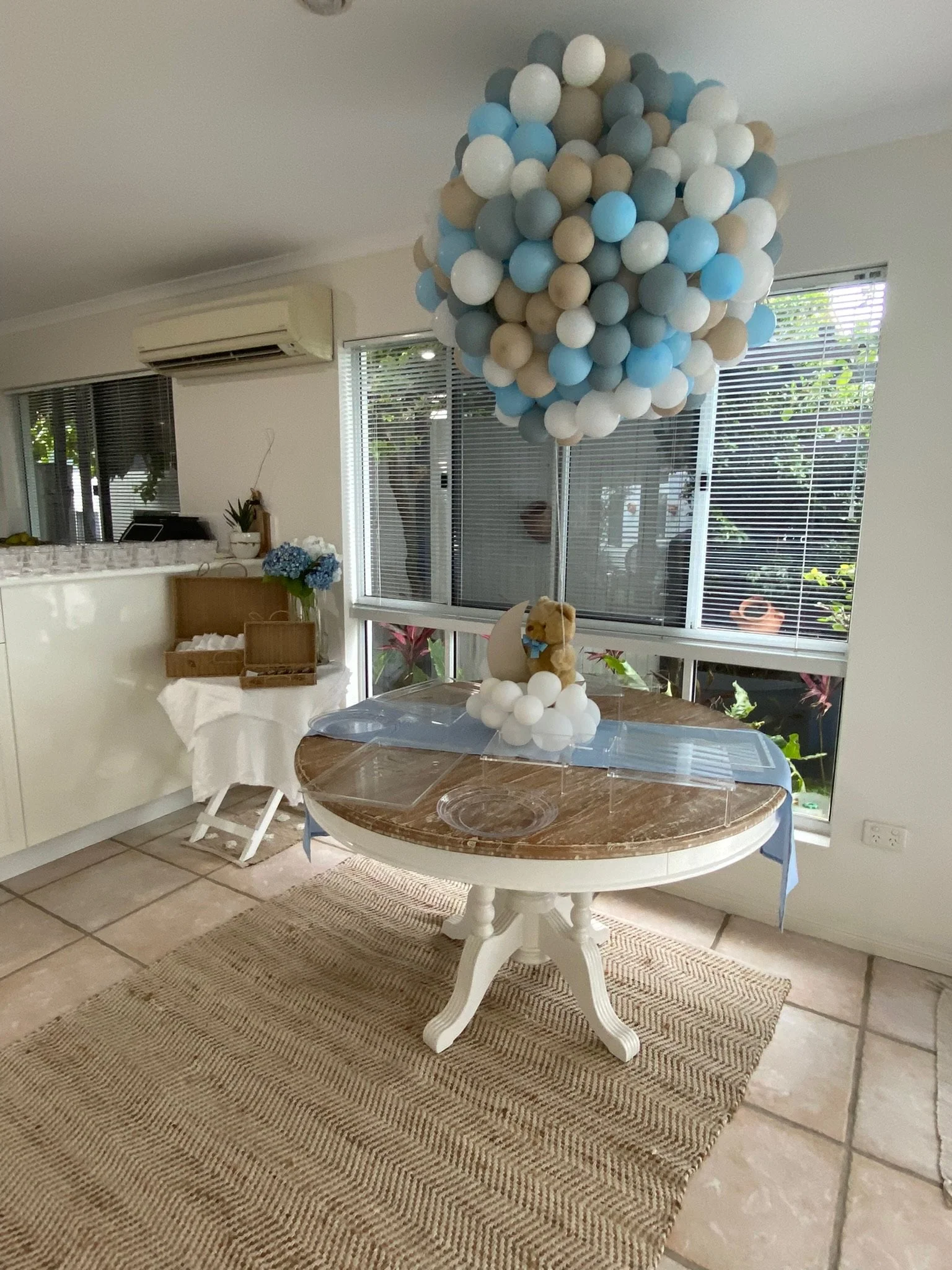 Baby shower decoration with a cluster of blue, white, and beige balloons hanging above a table, which has a teddy bear and more balloons on top, inside a bright room with large windows and tiled floor.