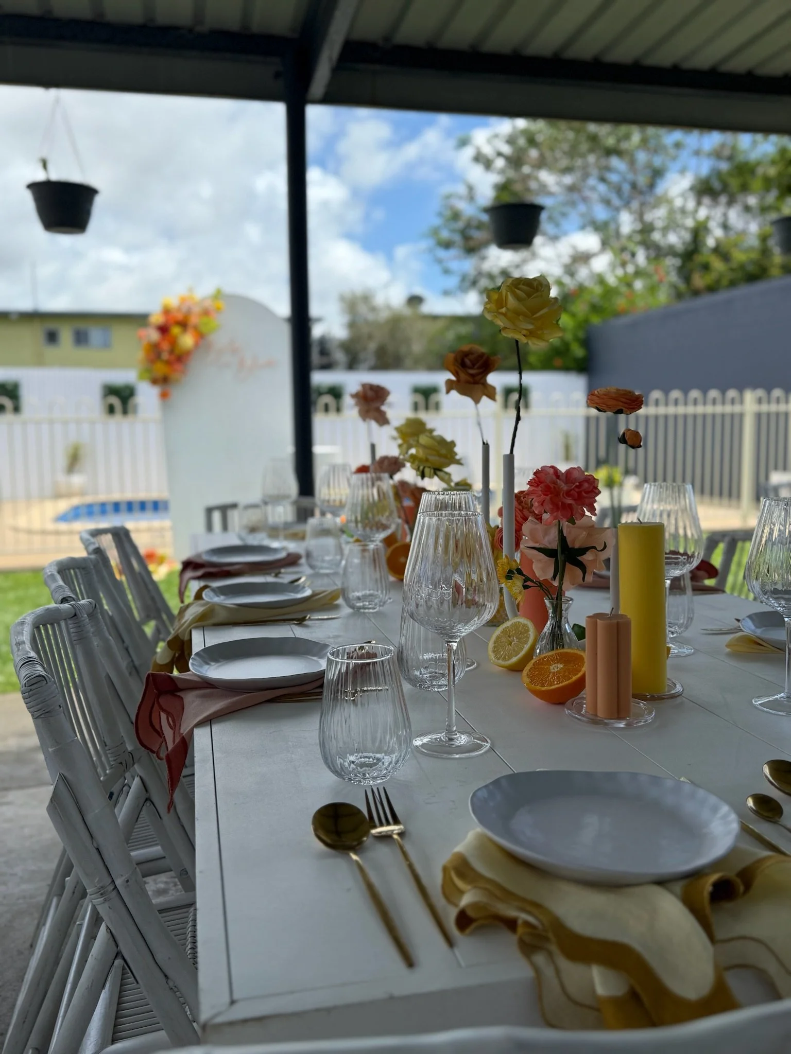 Decorated outdoor dining table with glassware, plates, cutlery, floral centerpieces, candles, and fruit slices on a covered patio with a backyard and cloudy sky in the background.