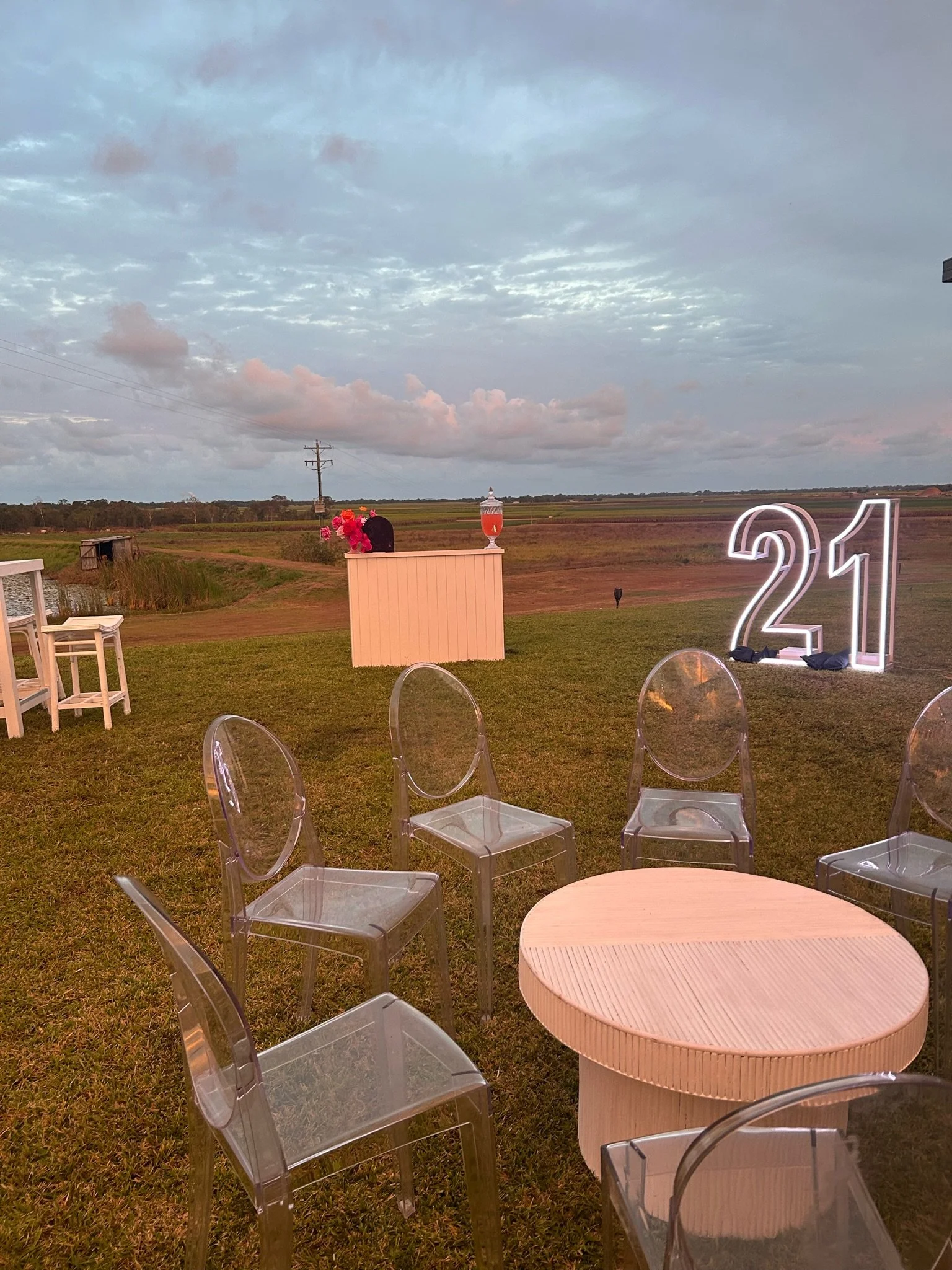 Outdoor celebration setup with transparent chairs around a round wooden table, a white backdrop with large illuminated numbers '21', a pink bar with flowers and a vase with a pink drink, set against a landscape with fields and a cloudy sky at sunset.