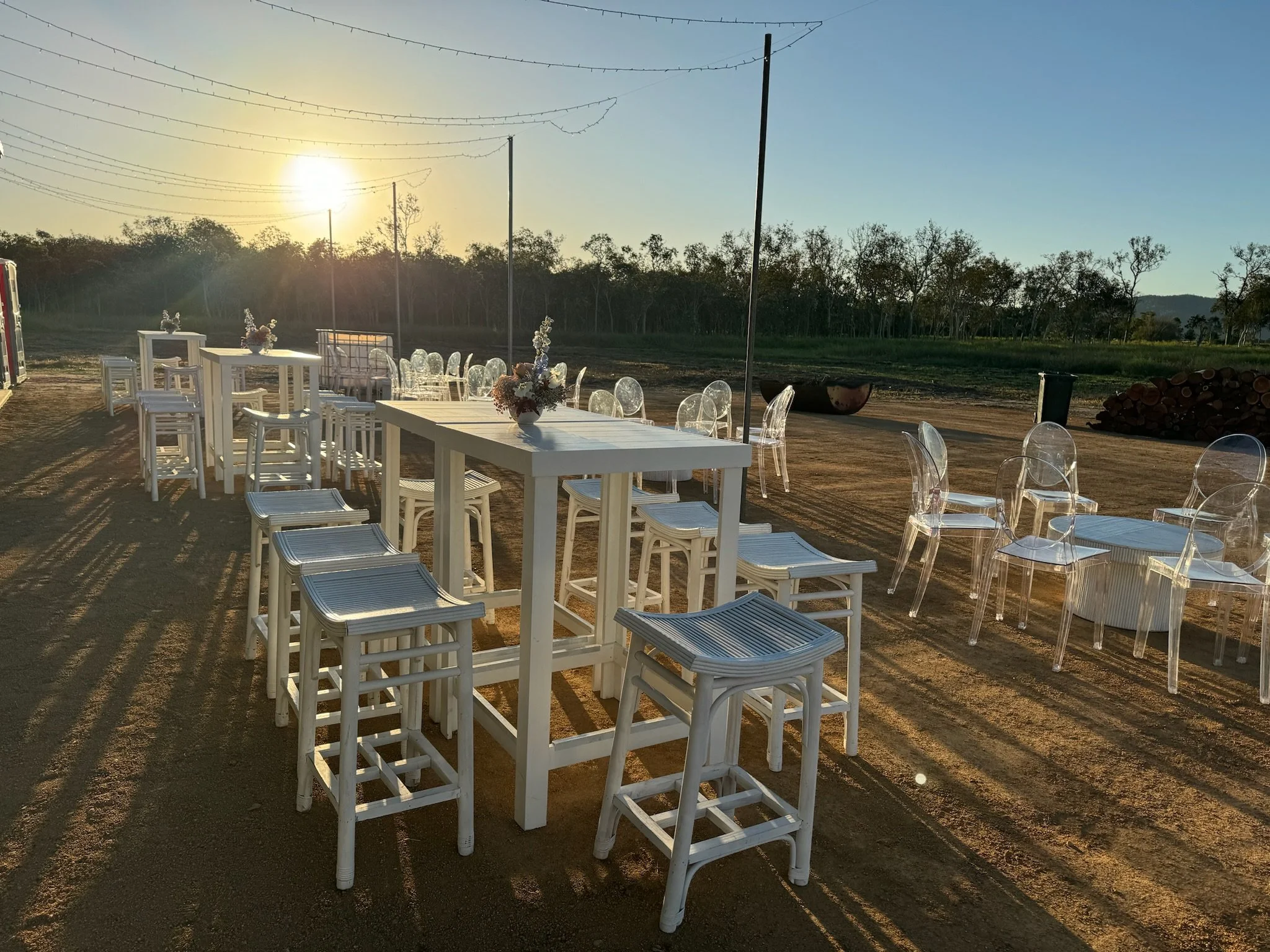 Outdoor event setup with white tables, white stools, and clear acrylic chairs on a dirt ground during sunset.