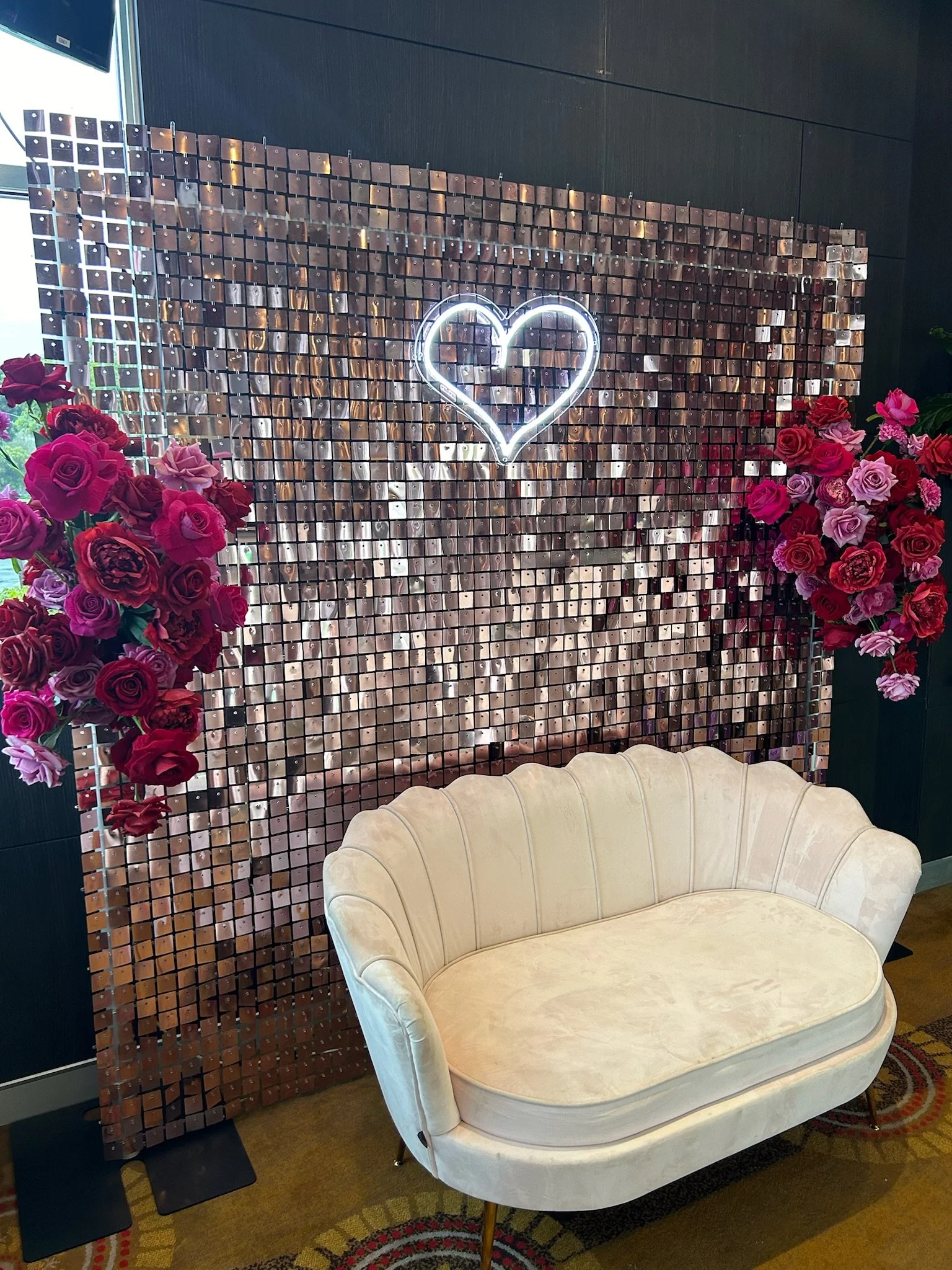 A decorative backdrop with a neon heart in the center, floral arrangements on either side, and a cream-colored vintage-style sofa in front.