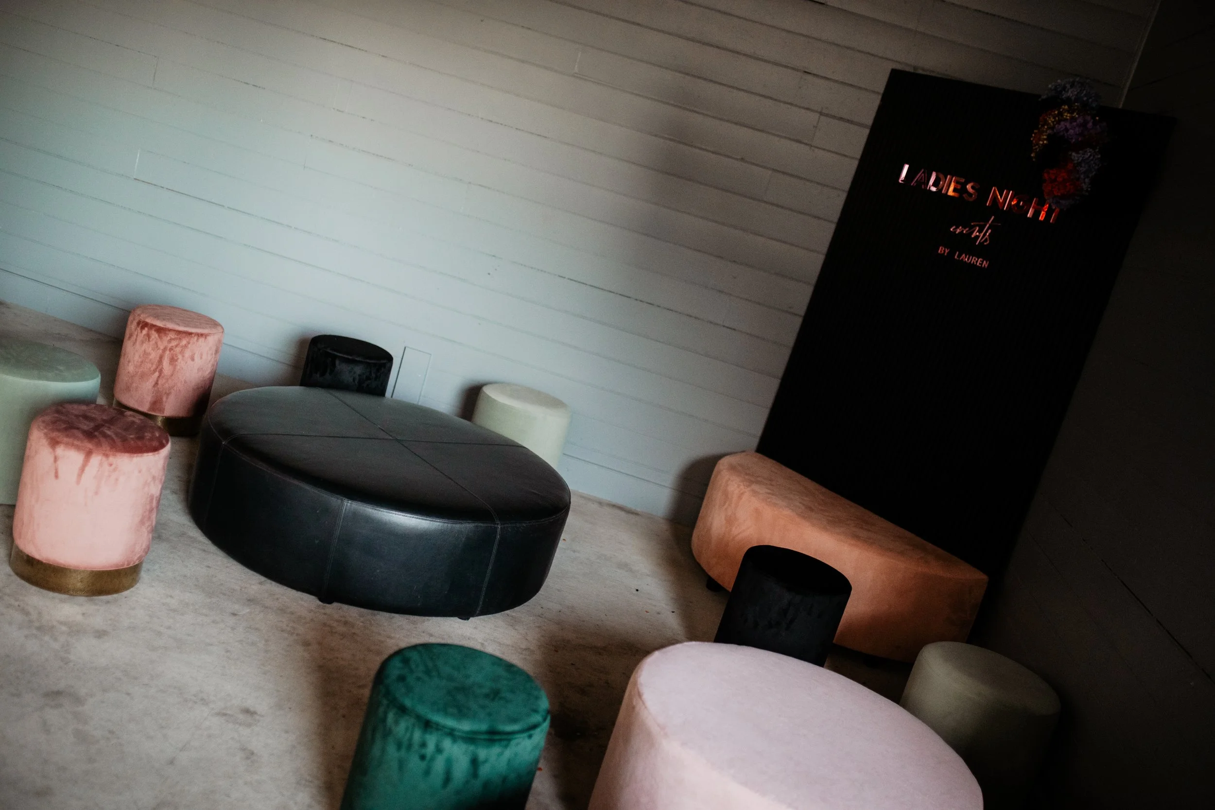 A lounge area with various velvet ottomans in pink, green, black, and cream, a large black leather ottoman, and a black sign with red and pink neon text reading 'LADIES NIGHT' and 'art by LAUREN'.