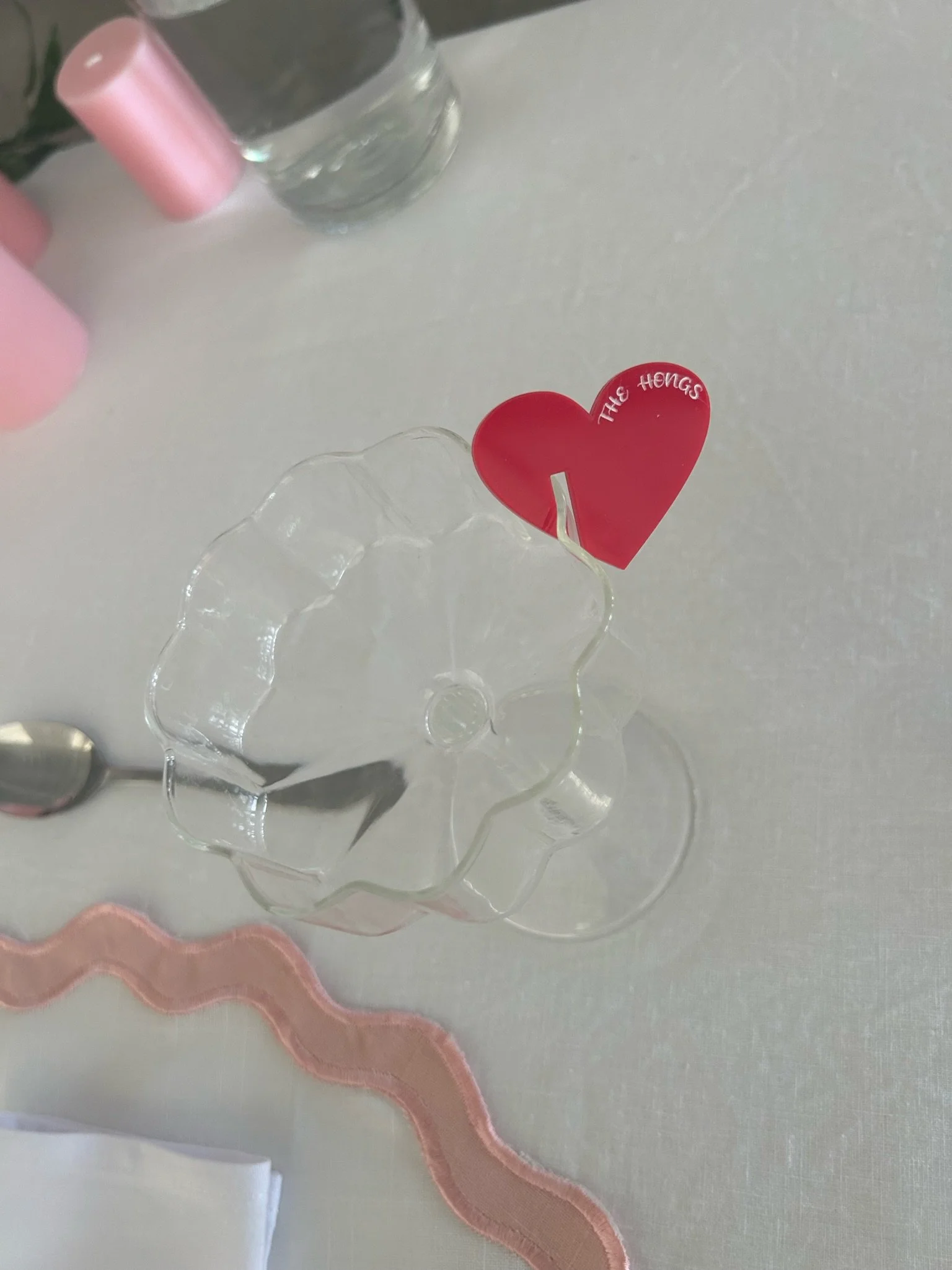 Clear glass dessert stand with a red heart-shaped note that reads 'The Hangs' attached to it, and a pink ribbon on a white table with a pink candle, a glass of water, a spoon, and a pink item in the background.