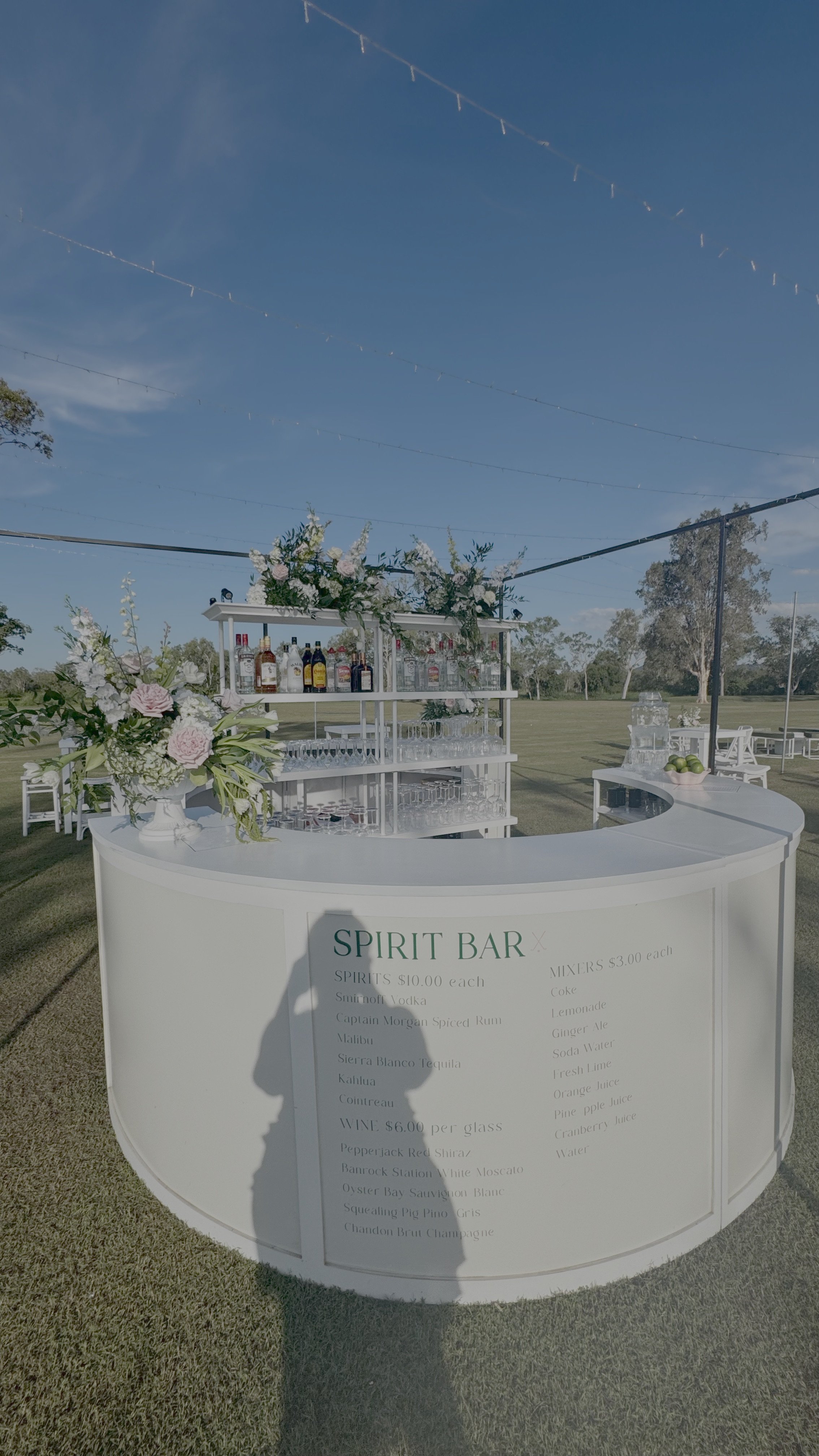 Outdoor white bar setup with floral arrangements, bottles of liquor, and glasses, labeled 'Spirit Bar', under a blue sky with string lights overhead.