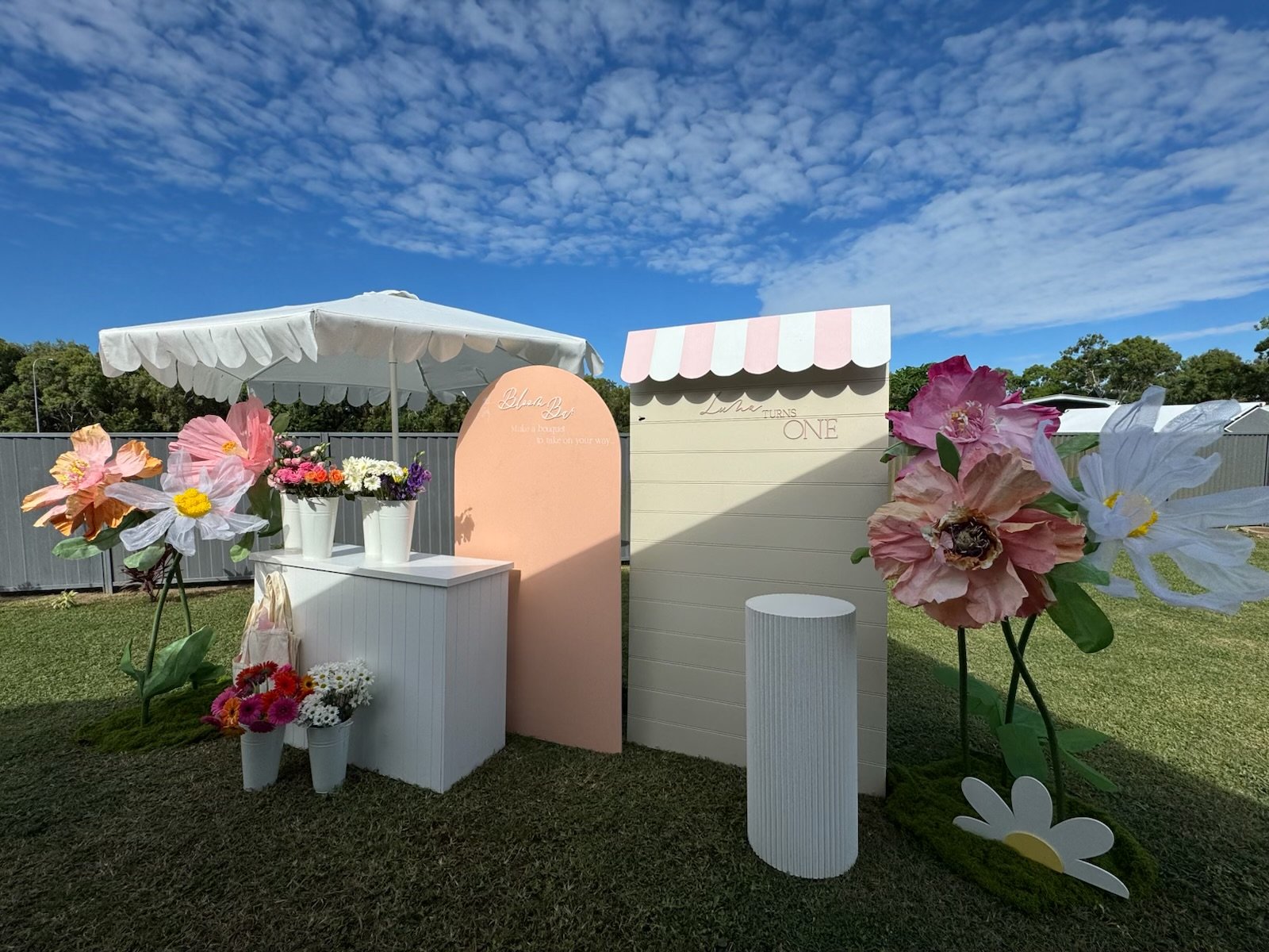 Decorative setup with large paper flowers, potted flowers, white and pink panels, and an umbrella outdoors on a sunny day.