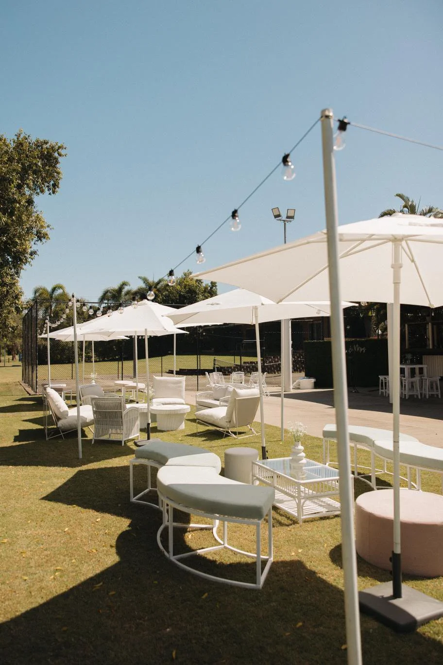 Outdoor seating area with white chairs, sofas, and tables under large white umbrellas, string lights overhead, on a sunny day with a clear blue sky.