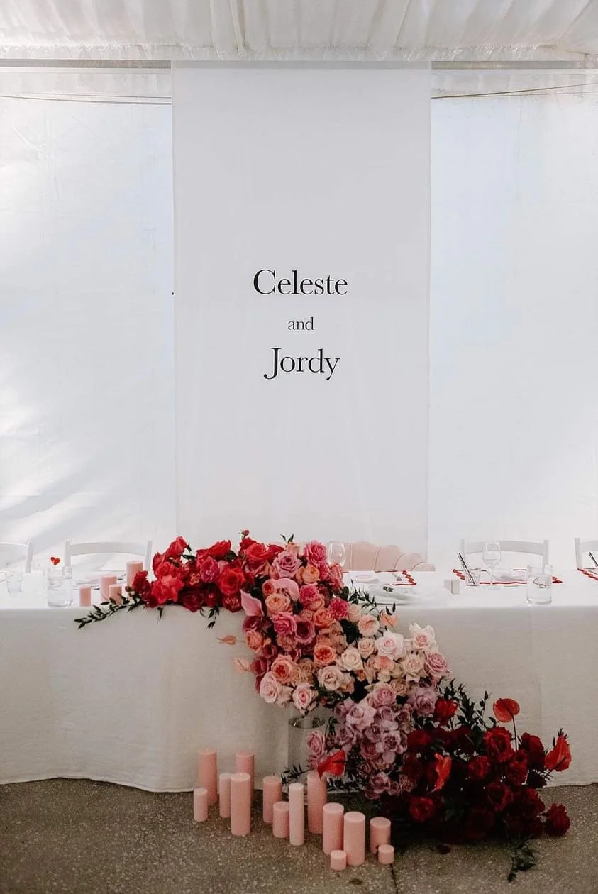 Wedding reception table with a large floral arrangement of pink, red, and peach roses. Candles arranged in front of the flowers. A white backdrop with a sign that reads 'Celeste and Jordy'.