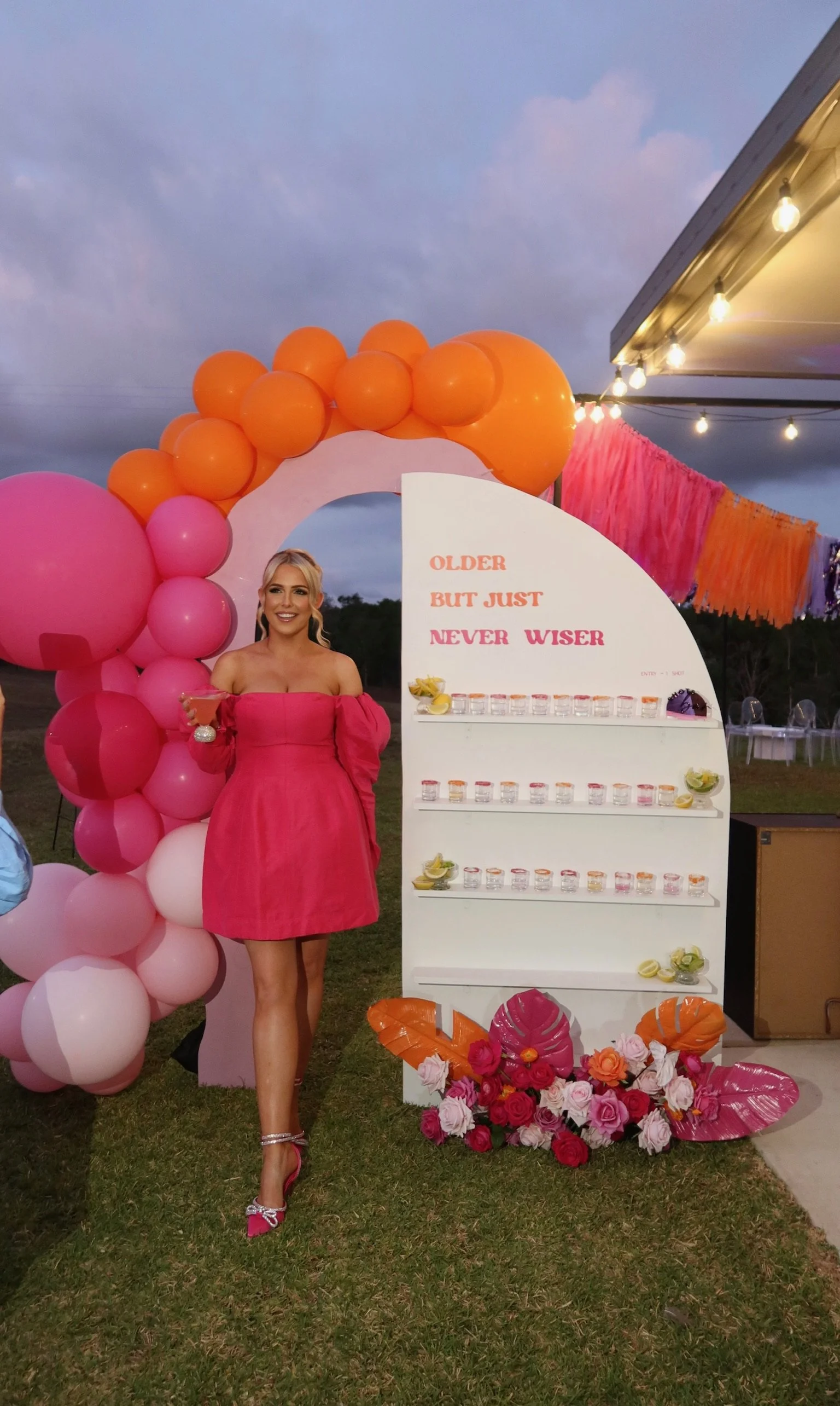 A woman in a pink off-shoulder dress holding a drink standing next to a decorative birthday display with balloons, flowers, and a sign that reads 'Older but just never wiser' at an outdoor celebration.
