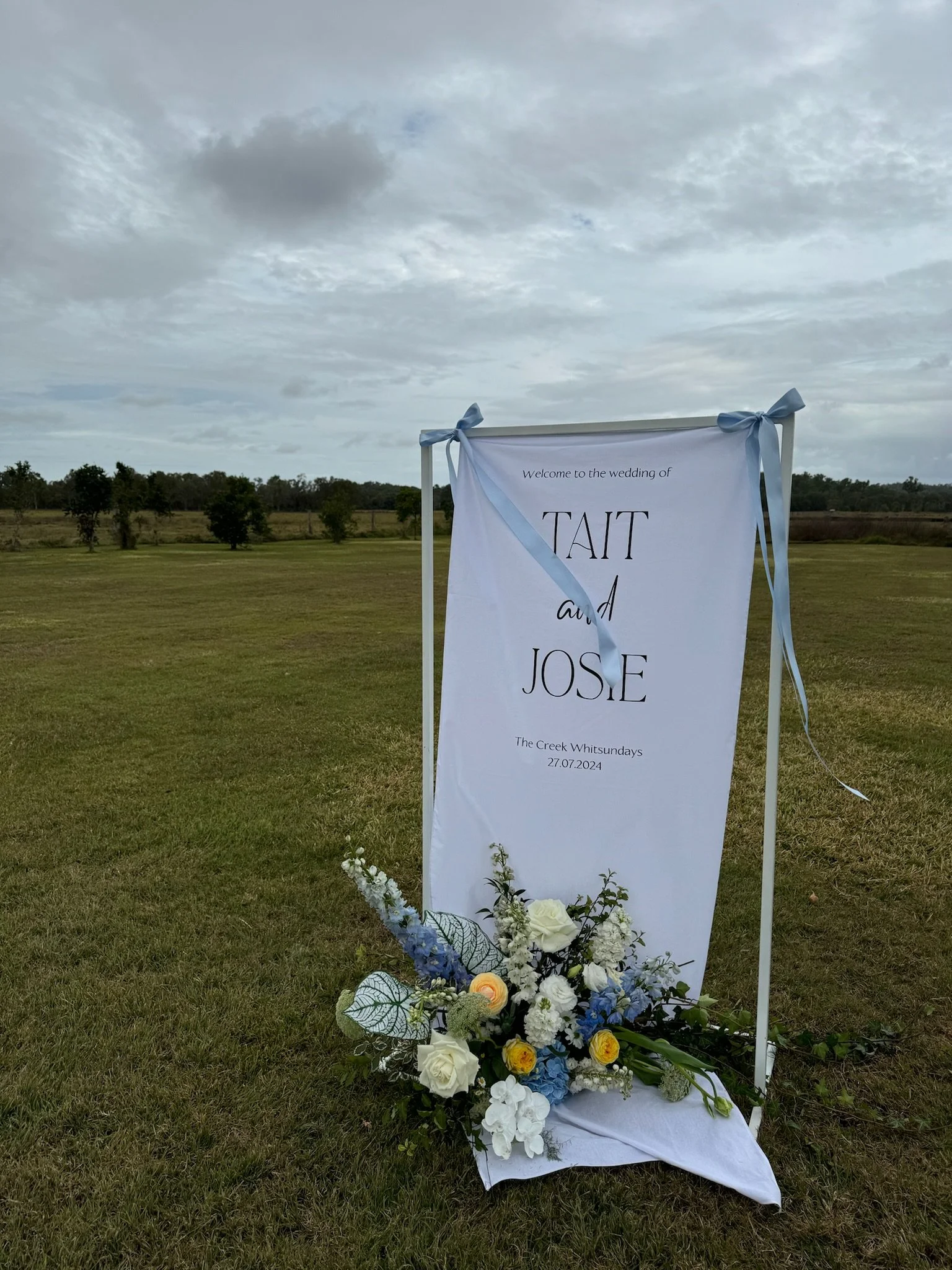 Wedding sign with floral decoration on grass, featuring white, yellow, and blue flowers with greenery, at an outdoor venue under cloudy sky.