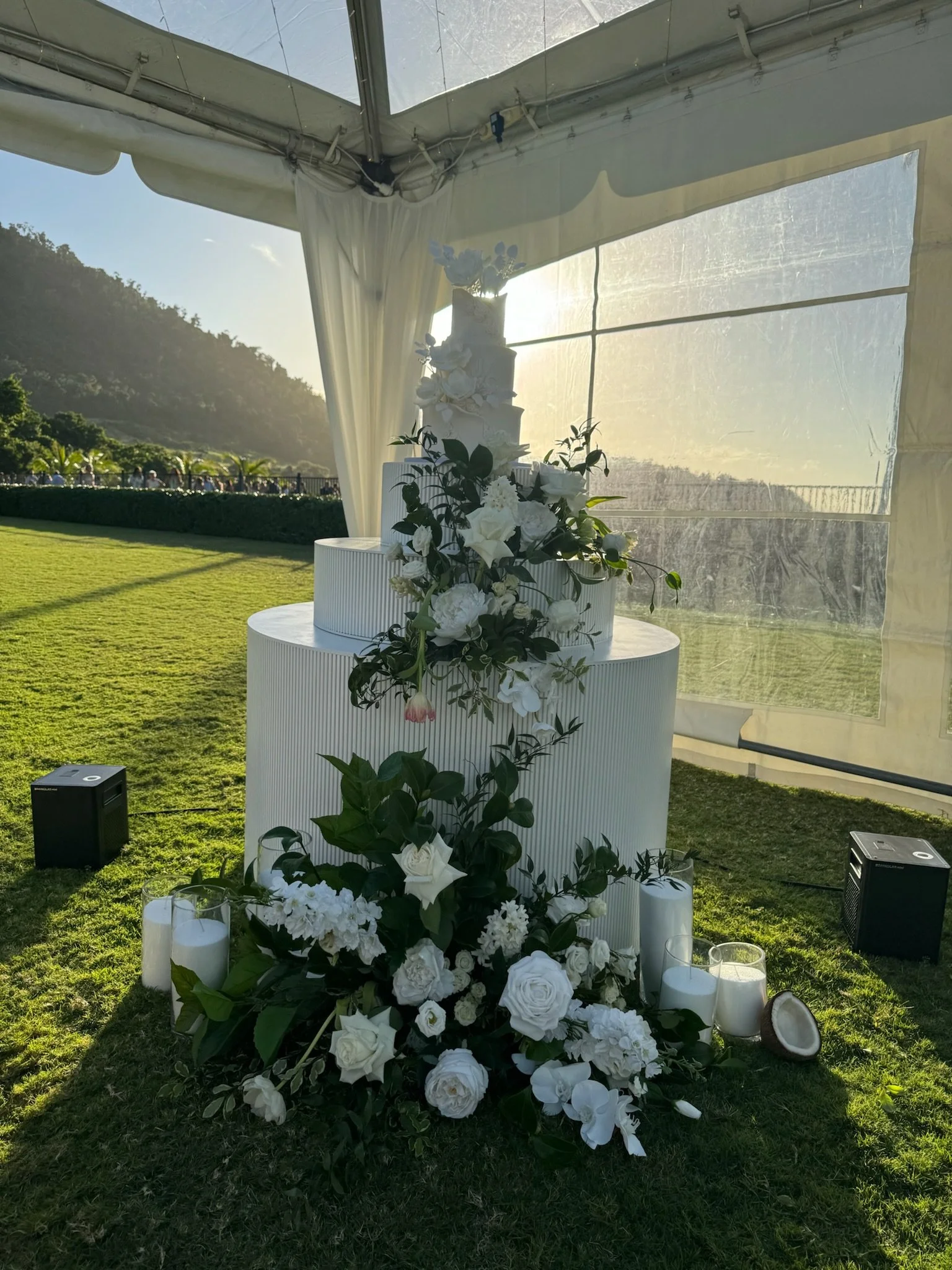 A white wedding cake decorated with white flowers, placed on a tiered stand, with a floral arrangement at the base, surrounded by candles, under a tent with a view of green grass and distant hills at sunset.