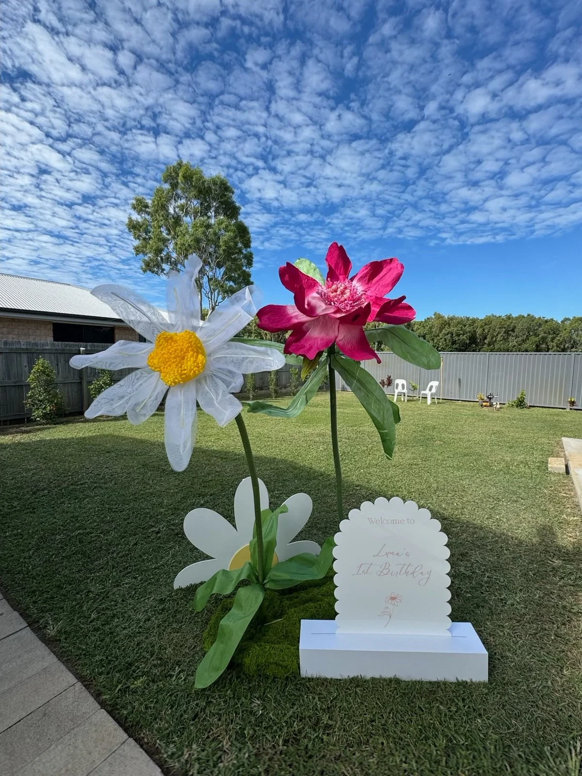 Decorative paper flowers, including a white daisy with a yellow center and a pink flower, placed outdoors on a grassy lawn with a cloudy blue sky, a tree, and a fence in the background. There is also a sign that says "Welcome to Lani's 1st Birthday."