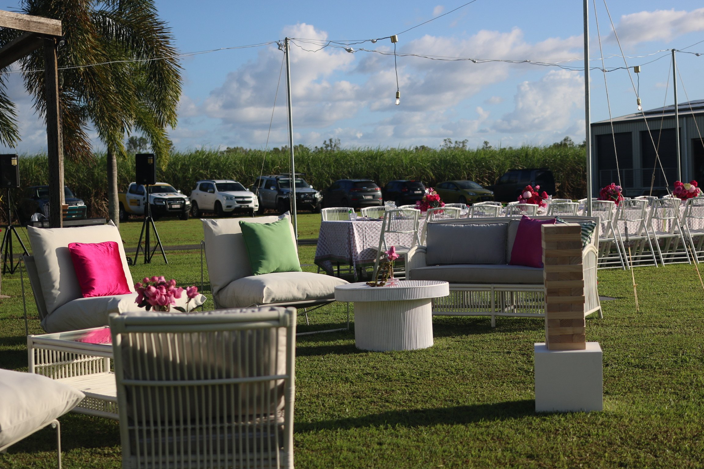 Outdoor event setup with white chairs, pink and green cushions, a white round table with flowers, and a lawn in the foreground. Cars parked in the background, palm trees, blue sky with clouds, and string lights overhead.