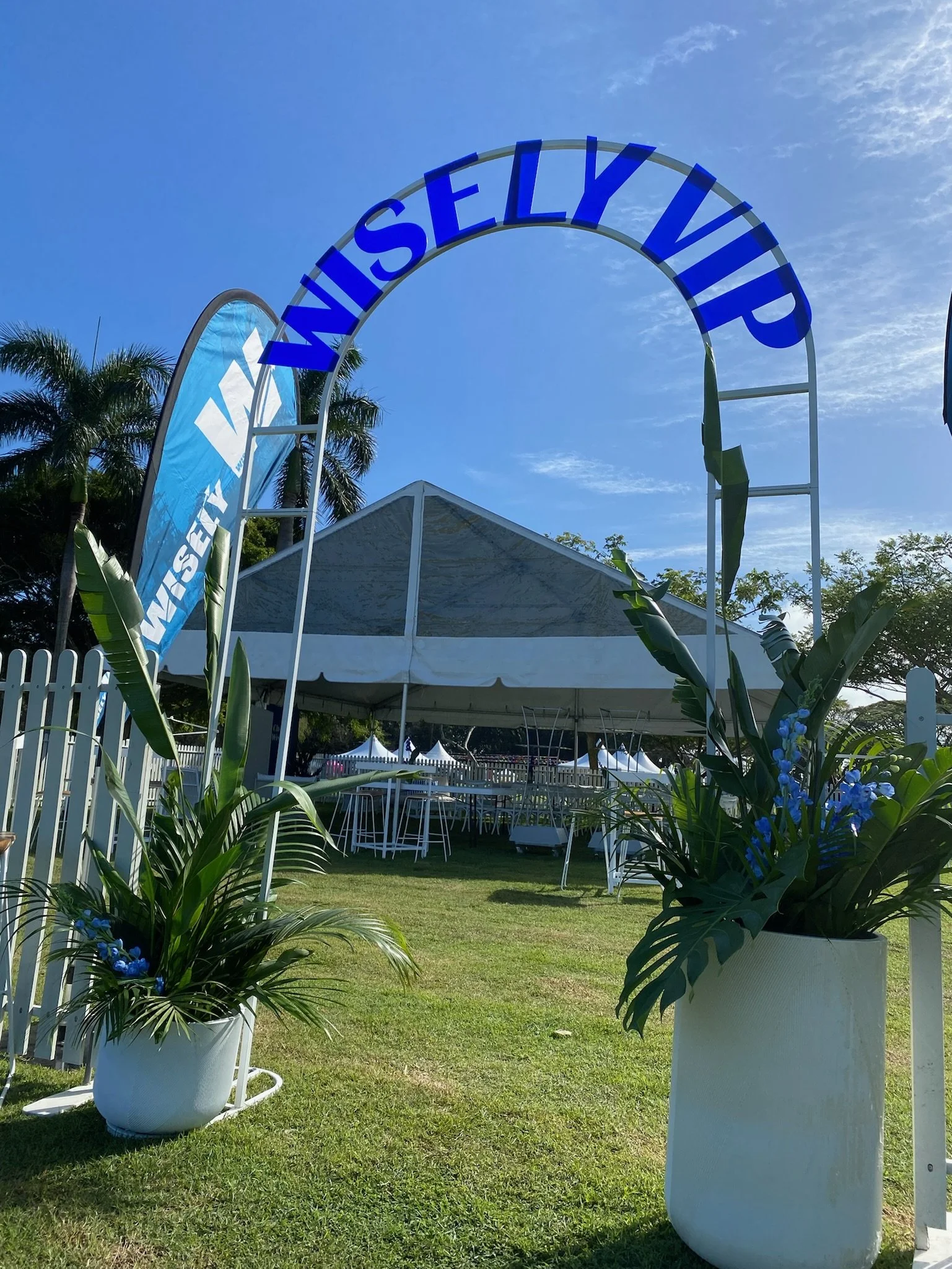 Outdoor event setup with a large white tent, white tables and chairs, decorative blue flowers in white pots, and a sign that reads "WISLEY VIP" under a clear blue sky.