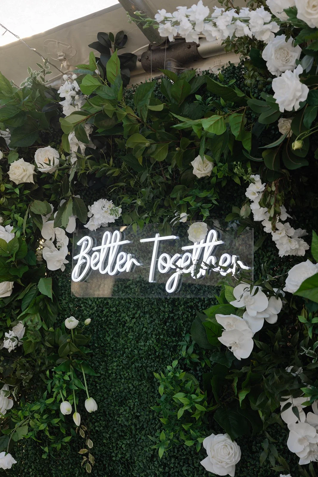 A floral wall with white roses and green leaves surrounding a neon sign that reads 'Better Together'.