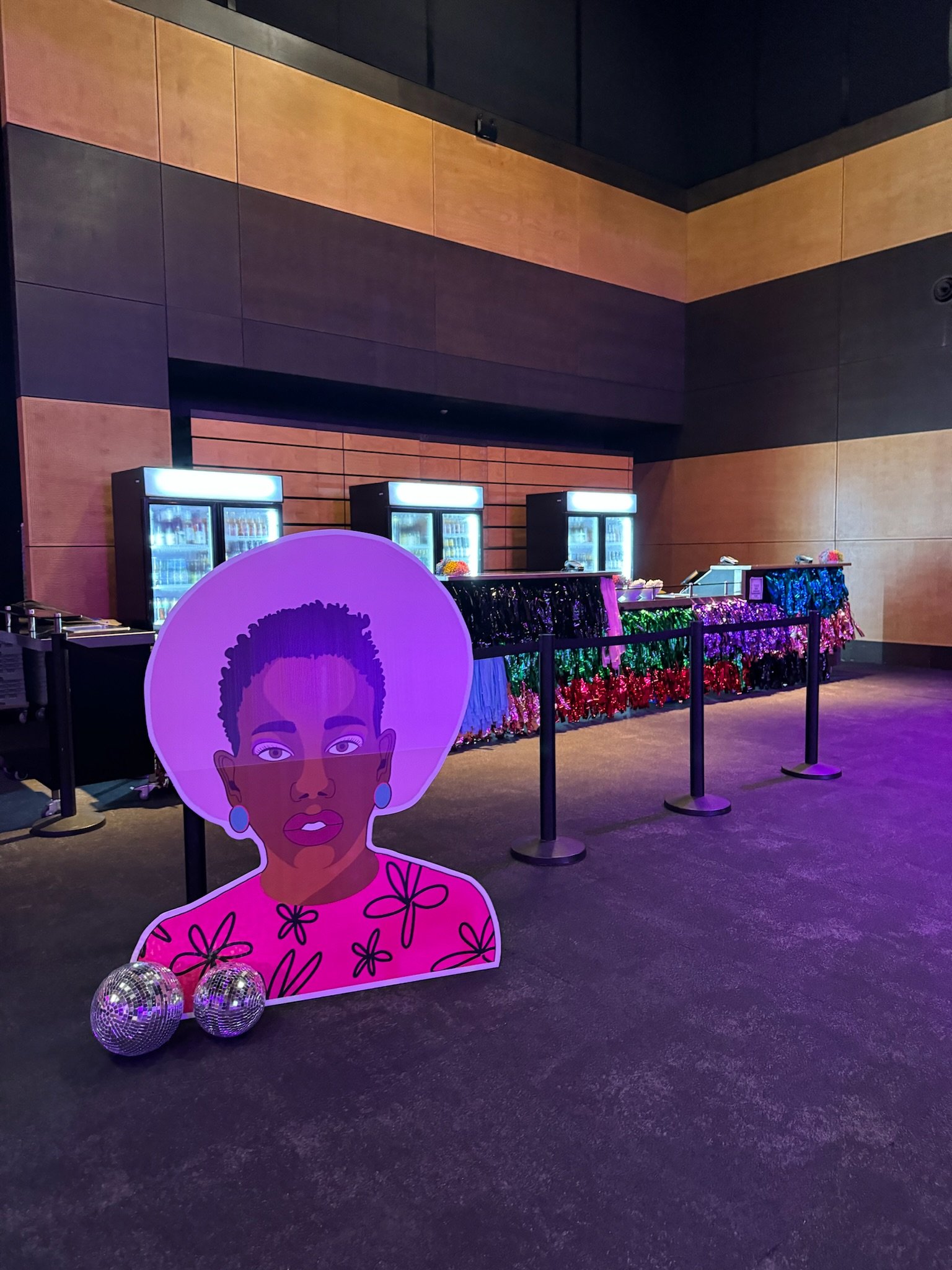 A colorful event setup with a cartoon cutout of a woman wearing a purple hat and pink dress with black floral designs, standing in front of a refreshment table decorated with multicolored tinsel and behind black stanchions. Two disco balls are placed