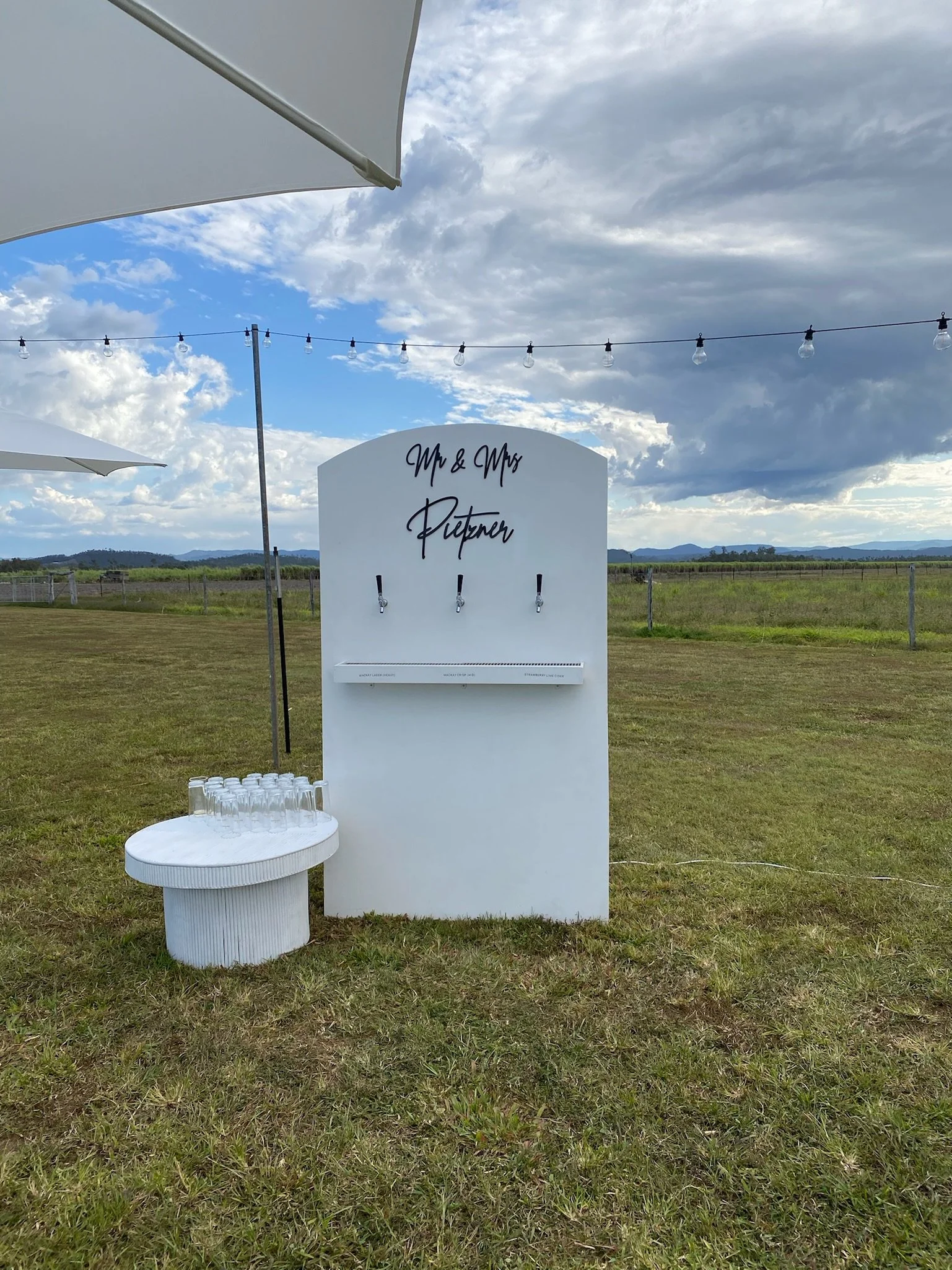 Outdoor wedding or event setup with a white drink station, beer taps, and glasses on a small round table, against a rural landscape with clouds and mountains in the background.