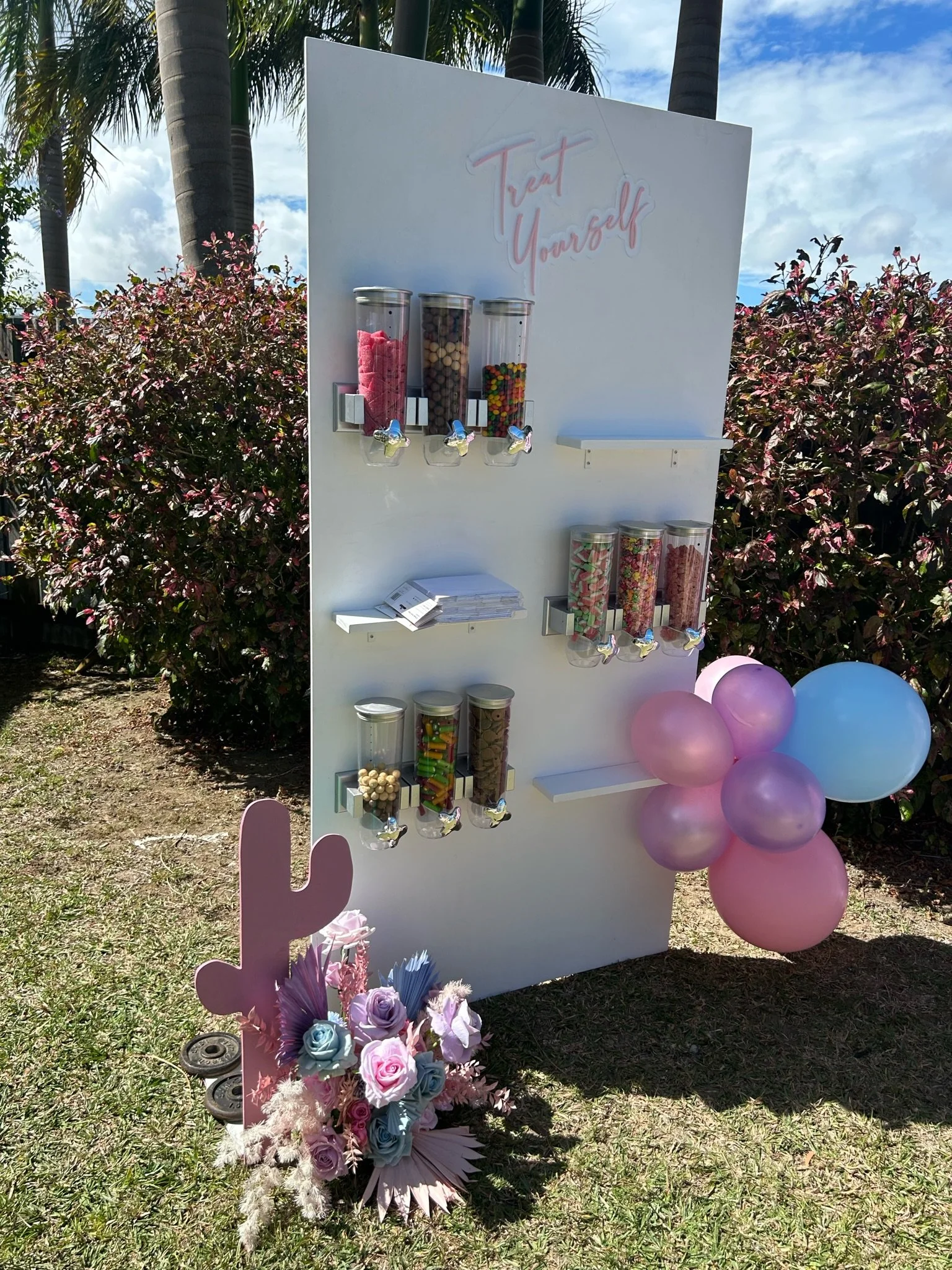 A DIY candy buffet station with a white display board featuring the phrase "Treat Yourself" in pink neon script. The board has three candy dispensers filled with pink marshmallows, brown, white, and colorful candies, with additional dispensers below 