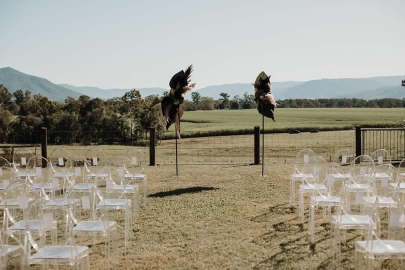 Outdoor event setup with transparent chairs arranged on grass, two decorative scarecrows, and open fields with mountains in the background.