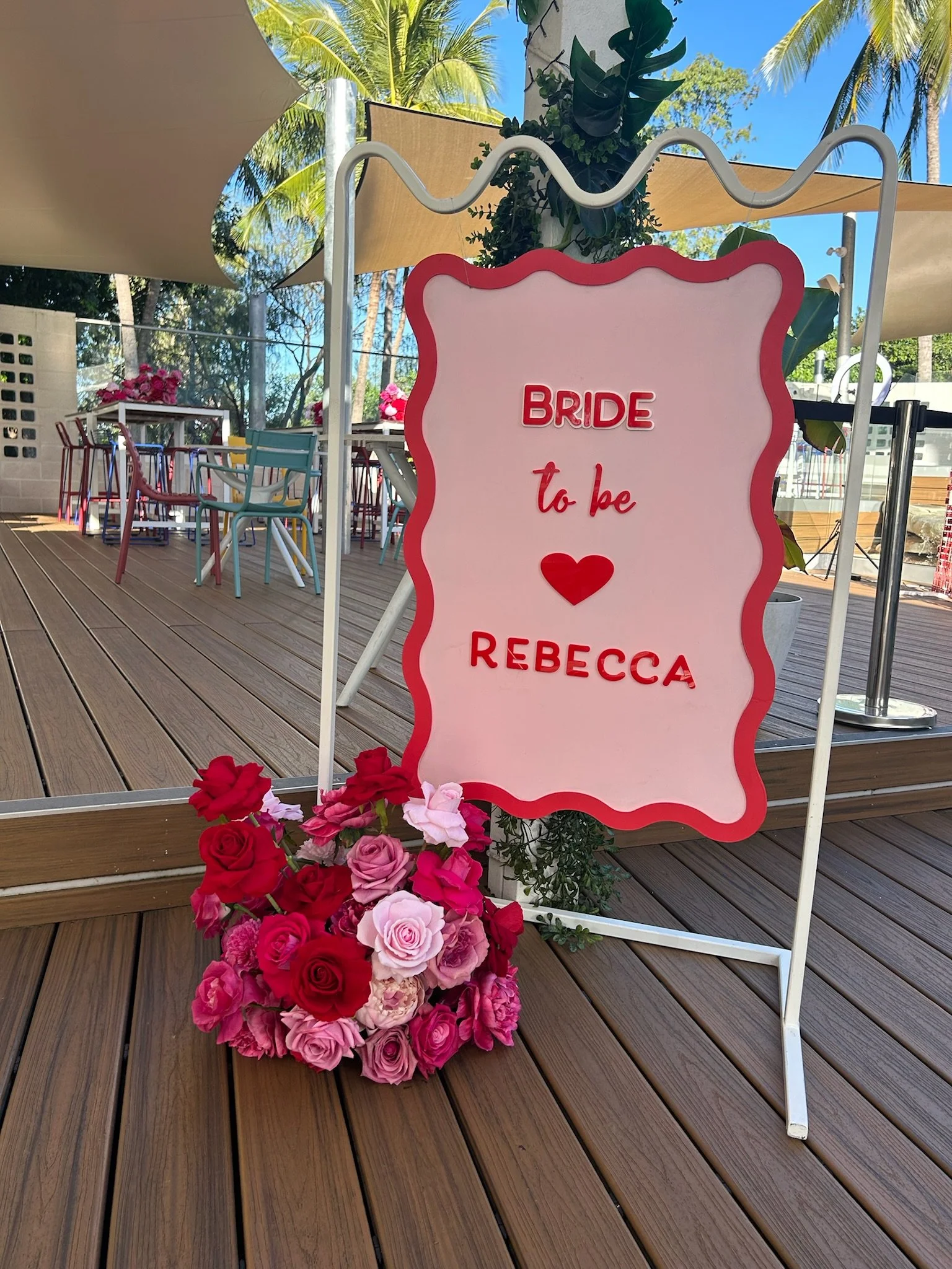 Sign that reads 'Bride to be Rebecca' surrounded by a floral arrangement of pink, red, and purple roses, set on a wooden deck with outdoor seating and tropical trees in the background.