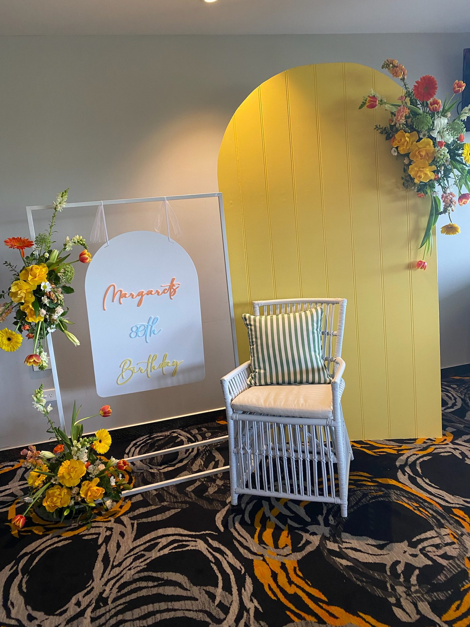 Decorative seating area with a white wicker chair and striped pillow, surrounded by yellow and floral arrangements, with a sign reading "Margaret's 8th Birthday" on a white frame and a yellow arched backdrop.