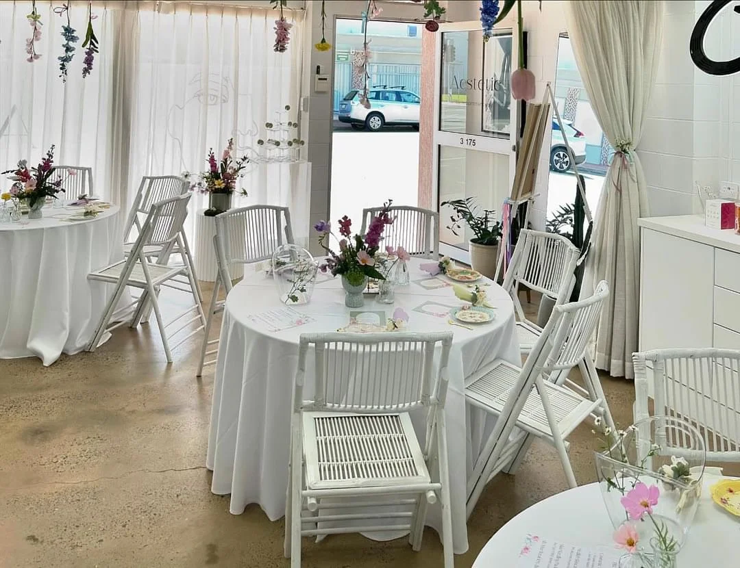 Interior of a bright, elegant event space decorated for a celebration, with white tables, chairs, floral centerpieces, hanging decorations, and a sunny front door.