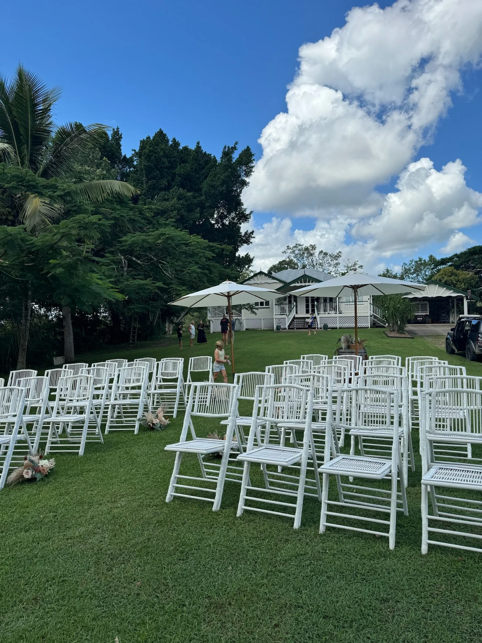 Arranged white chairs with floral decorations set on a green lawn for an outdoor event under umbrellas, with a house and trees in the background on a bright, partly cloudy day.
