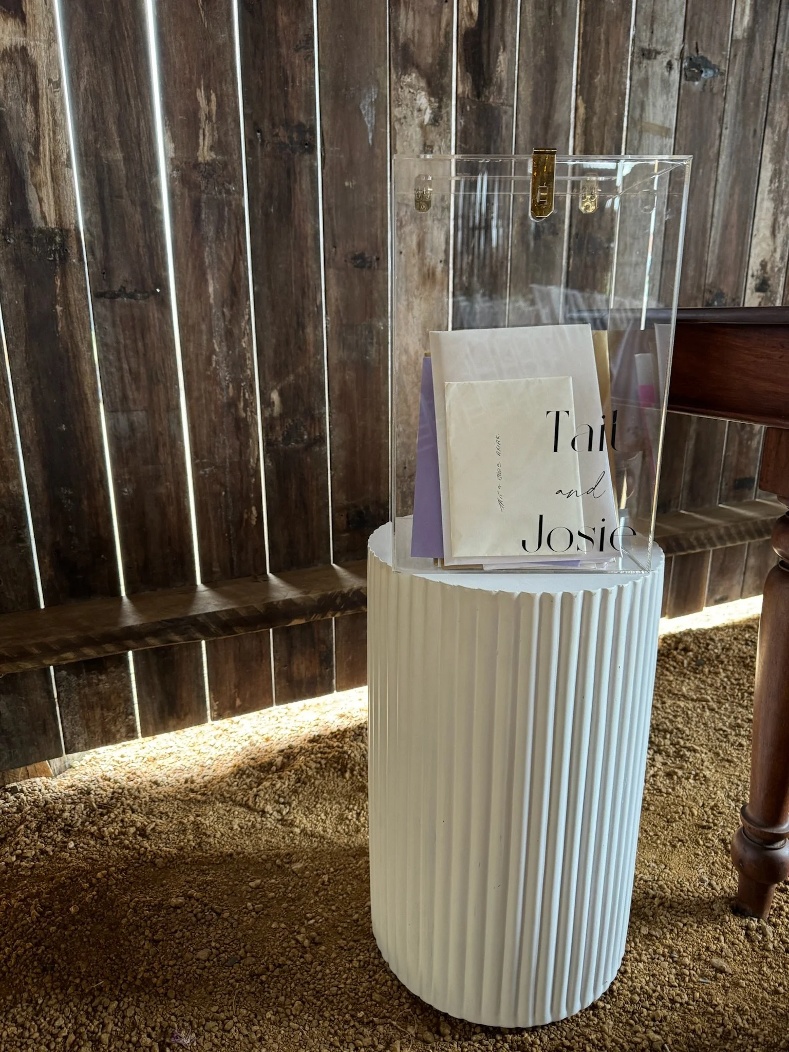 Clear acrylic box on a ribbed white pedestal holding paper wedding programs that read 'Tait and Josie'.