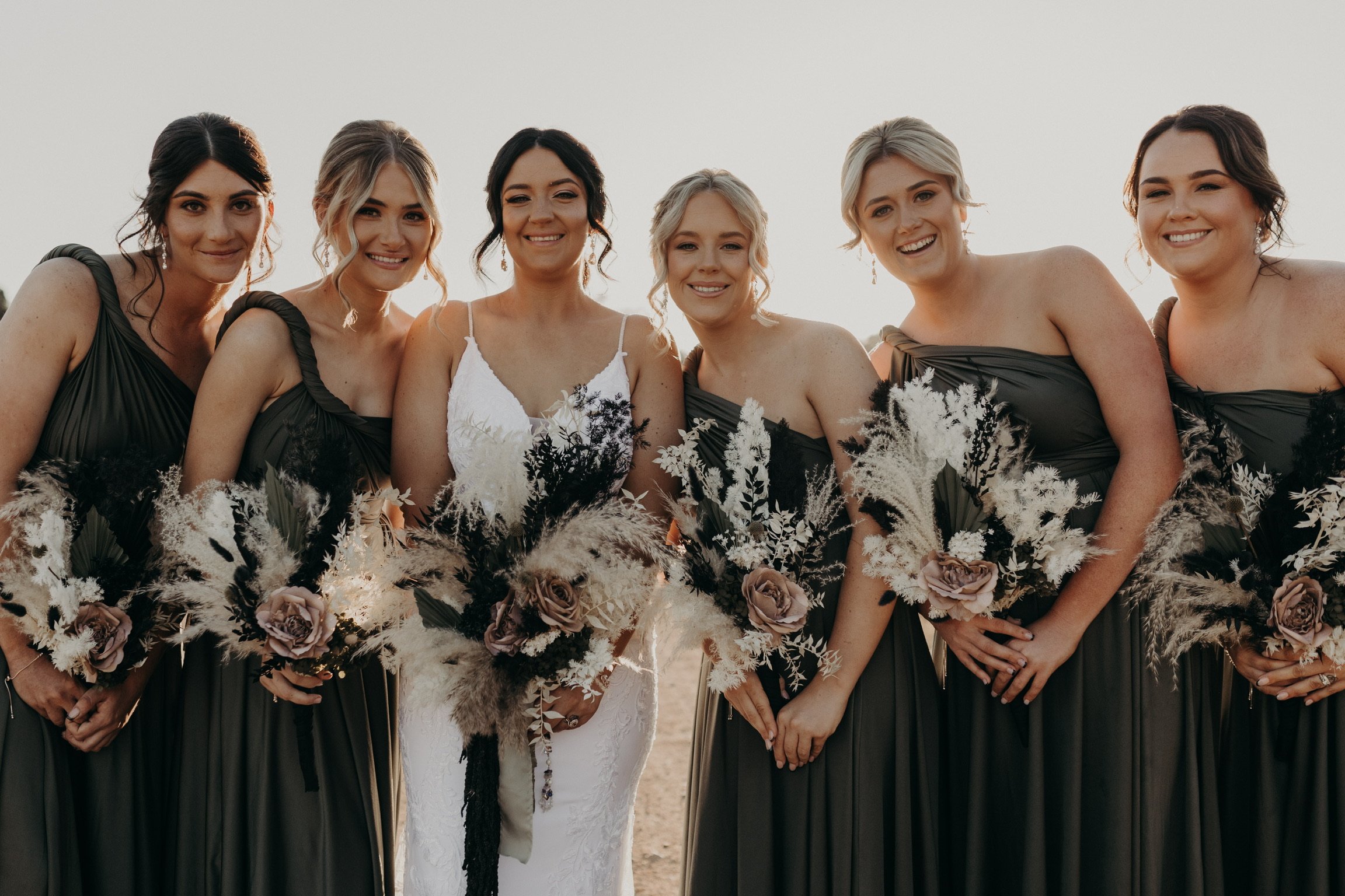 A bride and five bridesmaids standing outdoors, holding bouquets and smiling at the camera during a wedding.