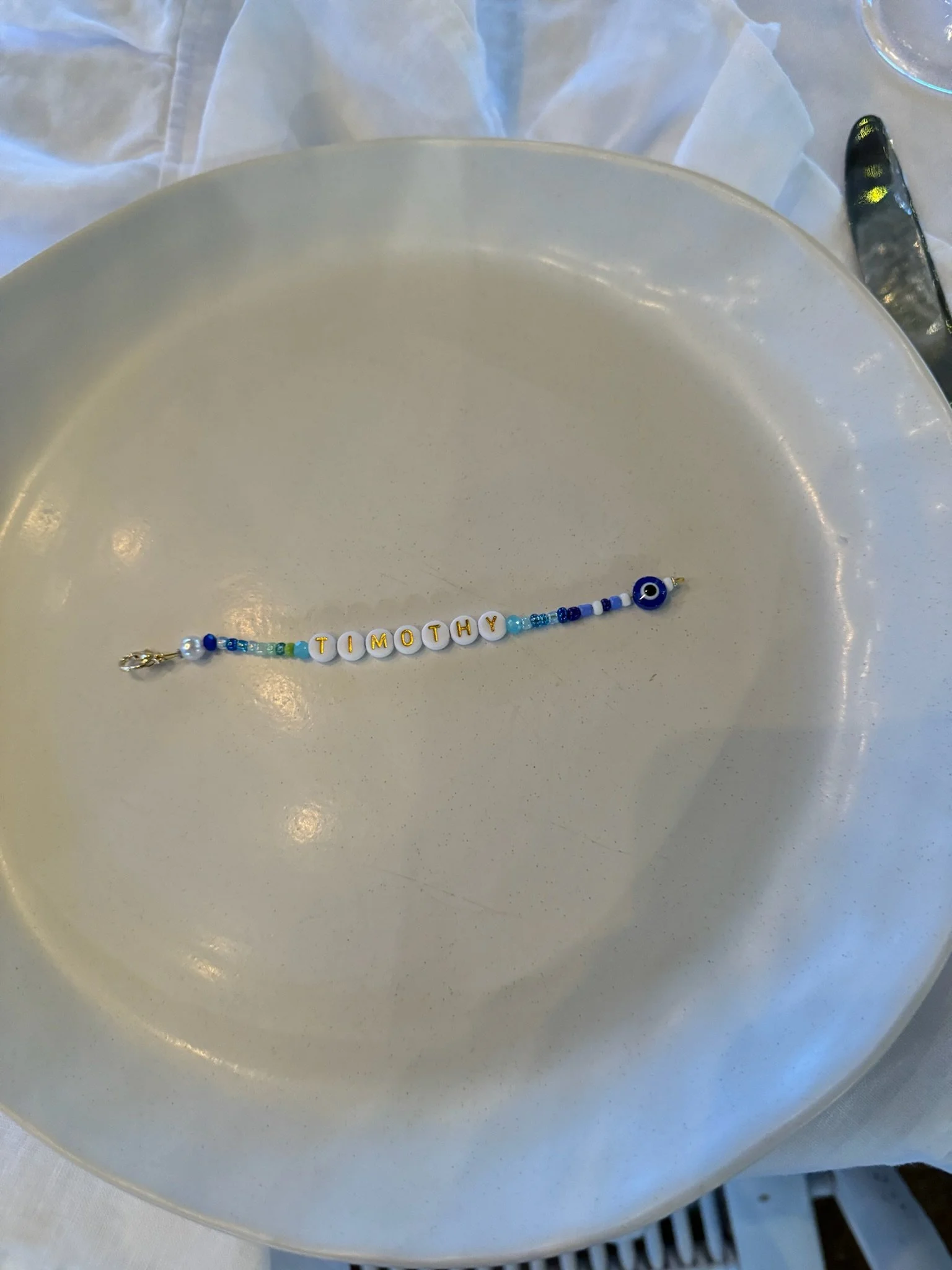 A beaded bracelet on a large white plate, spelling out the name 'Timothy' with white letter beads and decorated with blue and white beads and an evil eye charm.