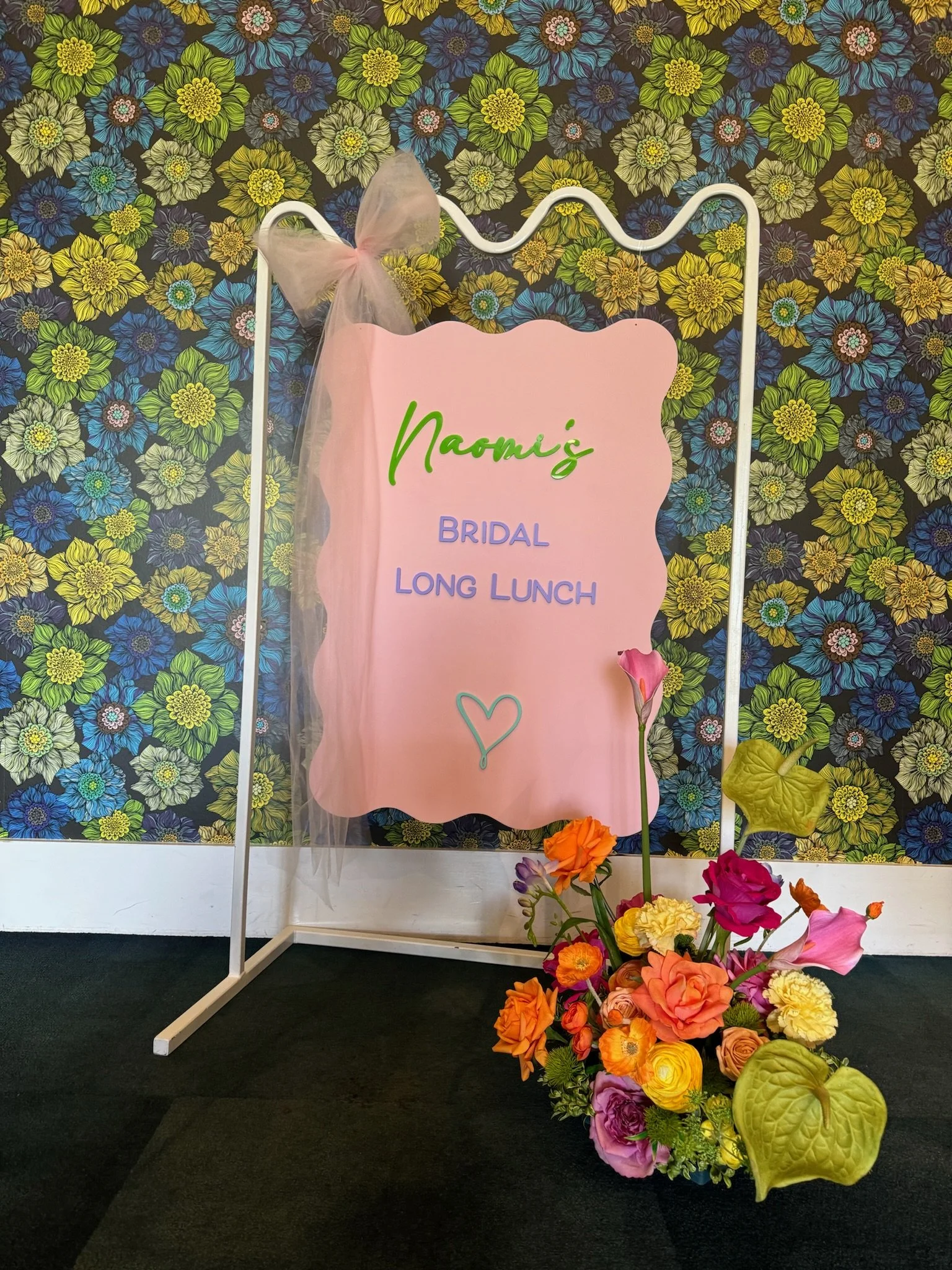 A floral display with a white easel holding a pink sign that reads "Naomi's Bridal Long Lunch" in colorful text, decorated with a pink tulle bow, and a vibrant bouquet of flowers at the base, against a background of yellow, green, blue, and purple fl
