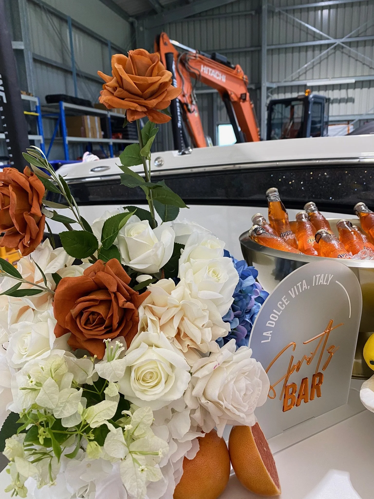 A bouquet of roses and flowers with white, orange, and blue blossoms, placed near a bowl of orange glass soda bottles on a counter inside a warehouse or garage with construction equipment.