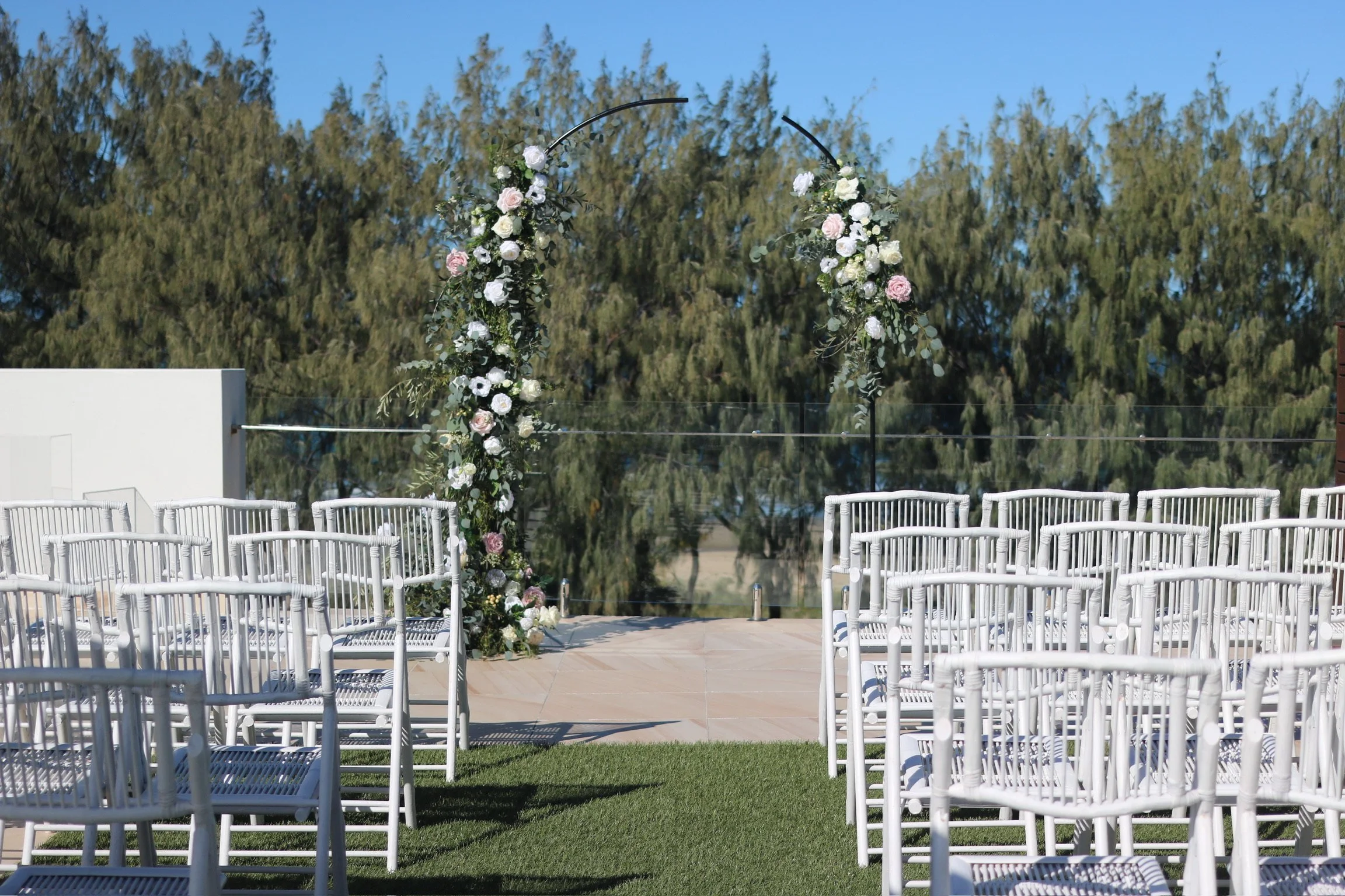 Outdoor wedding setup with white chairs facing a floral arch on a grassy area under clear skies.