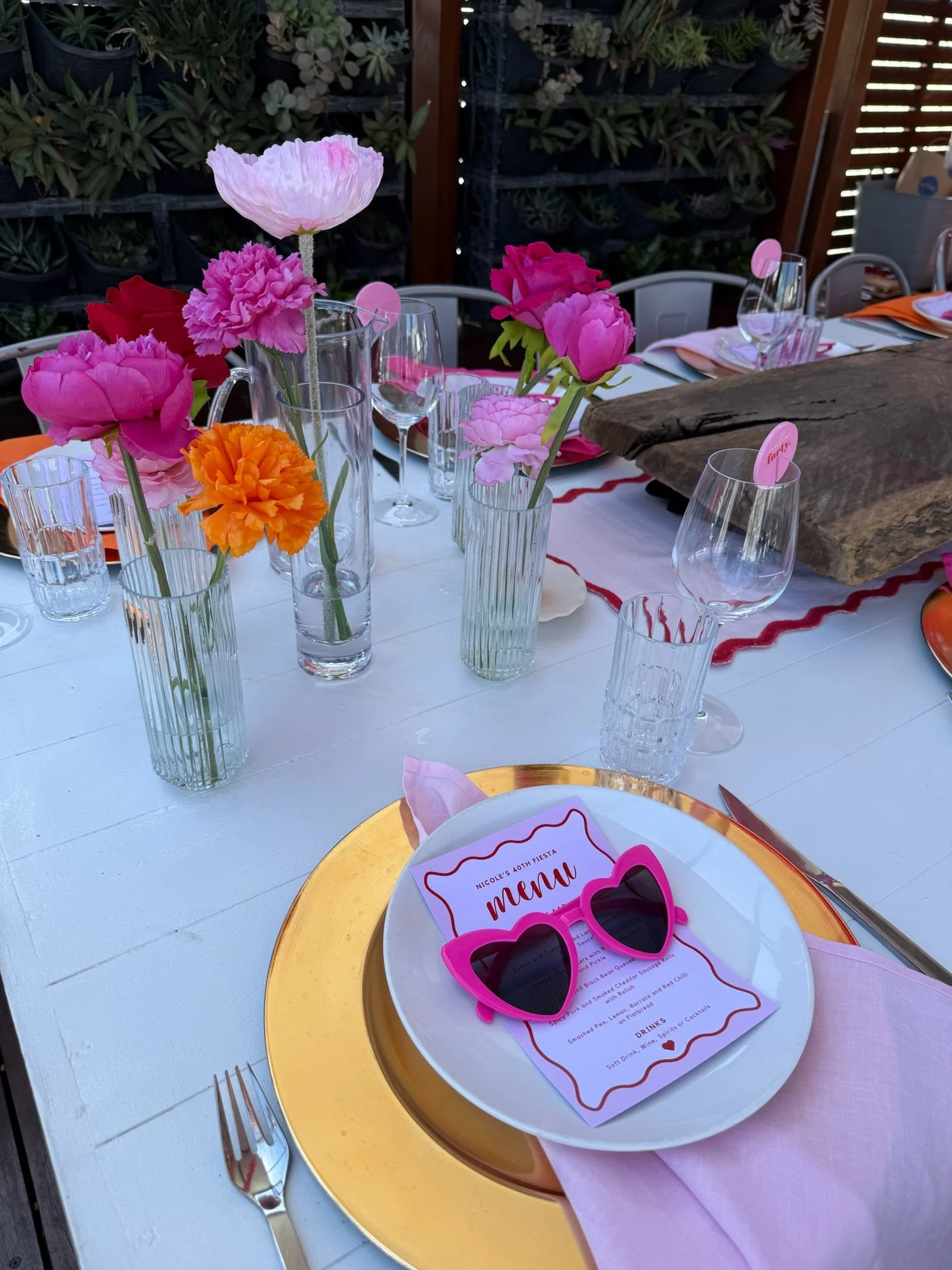 Decorated outdoor table with pink, orange, and purple flowers in tall glass vases, set with white plates, gold charger plates, pink sunglasses, and a themed menu for a birthday celebration. The table has pink napkins, glassware, and a rustic wooden c